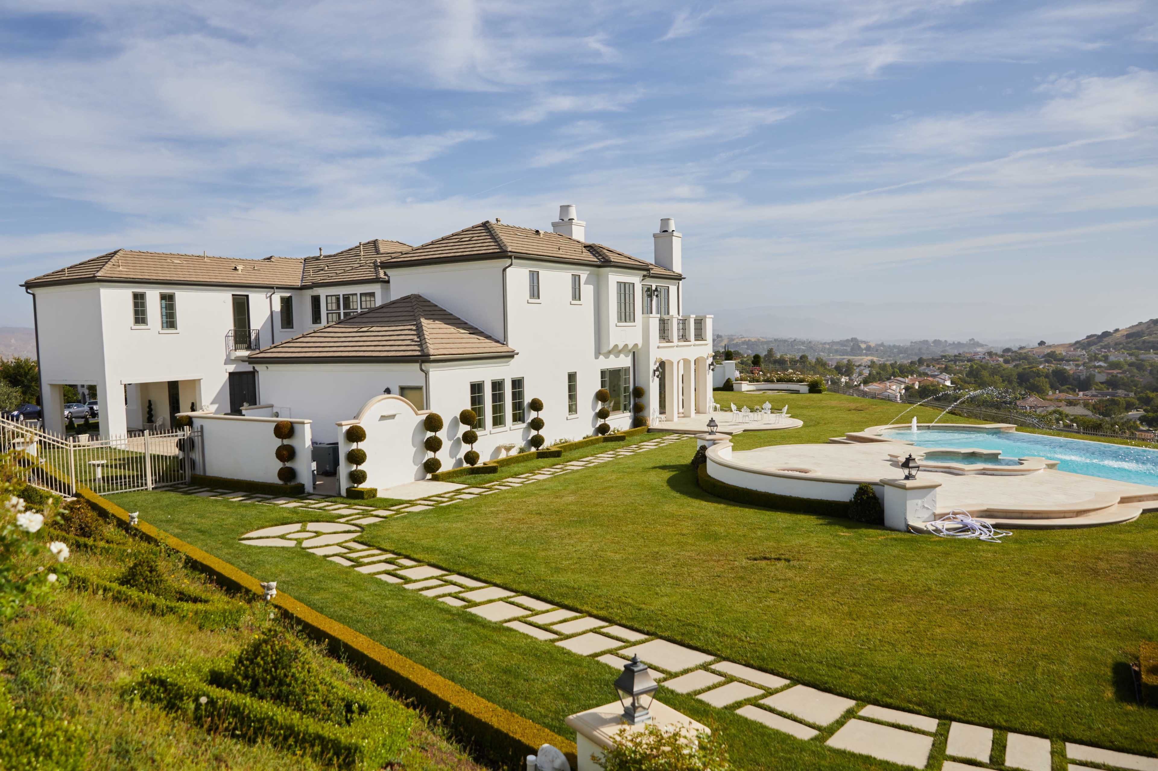A large white mansion with a manicured lawn and a swimming pool is situated on a hillside, featuring a stone pathway leading to the house.