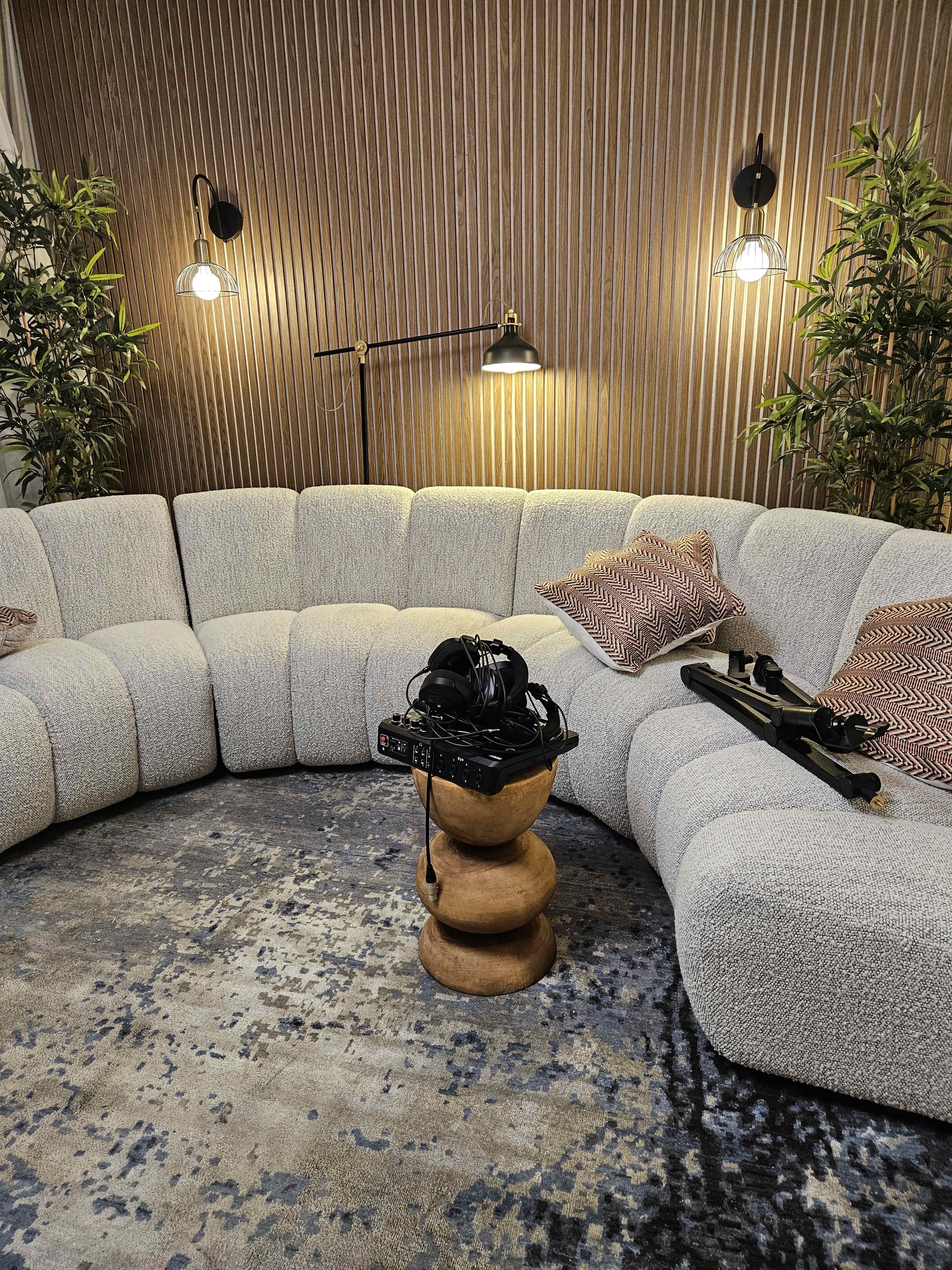 A modern, curved couch sits in a cozy room with wall-mounted lamps and decorative plants, accompanied by a wooden stool holding headphones and a microphone.