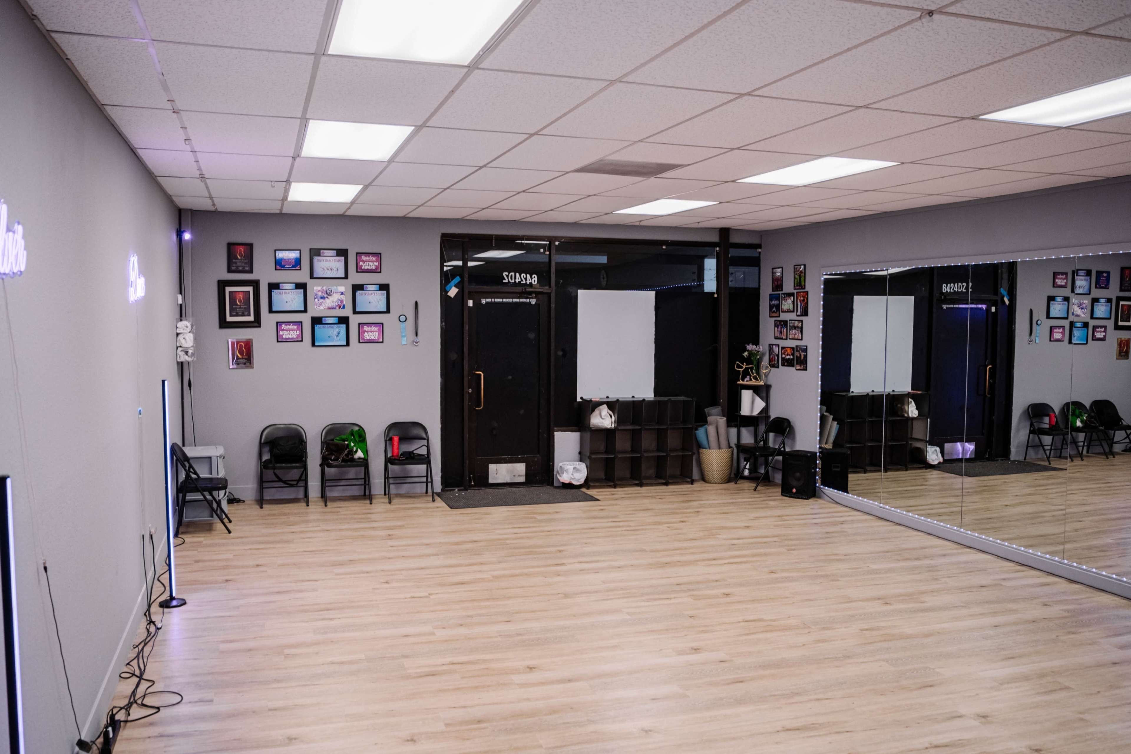 The image shows a spacious dance studio with wooden flooring, a large mirror on one wall, and several black chairs arranged along the opposite wall.