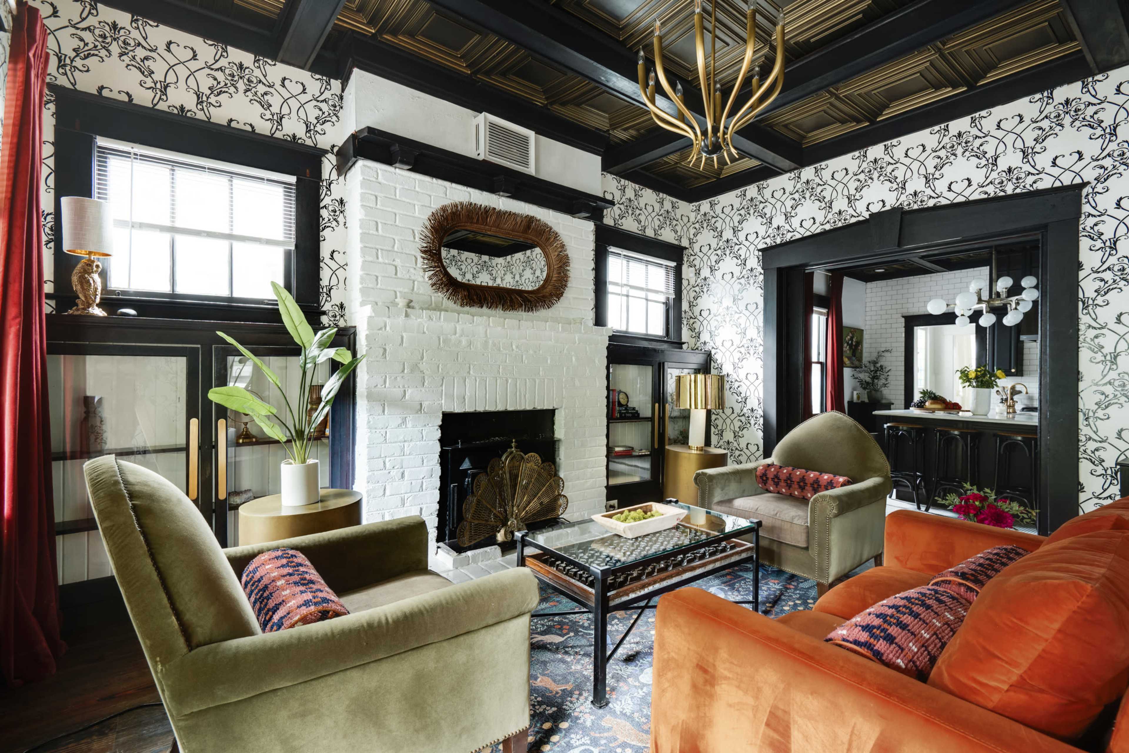 The image depicts a stylish living room featuring a white brick fireplace, a mix of upholstered seating in green and orange, and patterned wallpaper.