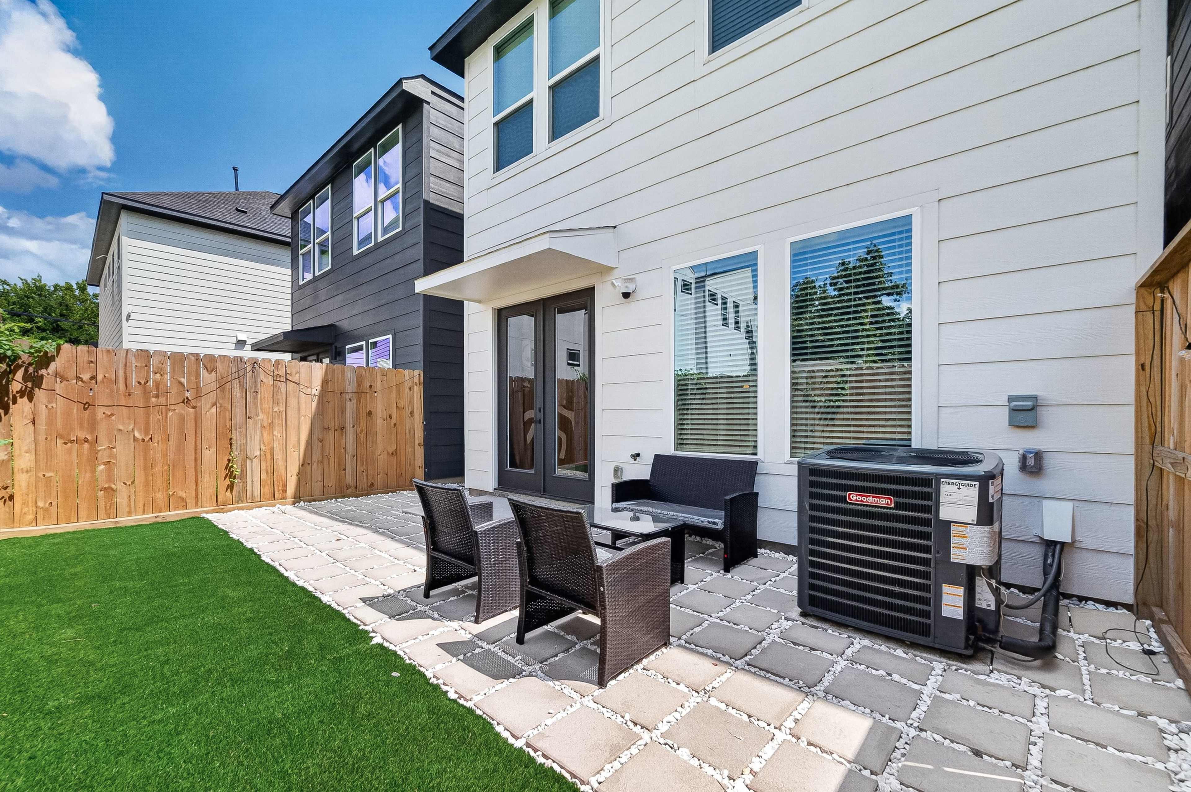 The image shows a modern backyard patio area with a seating arrangement, an air conditioning unit, and a wooden fence.