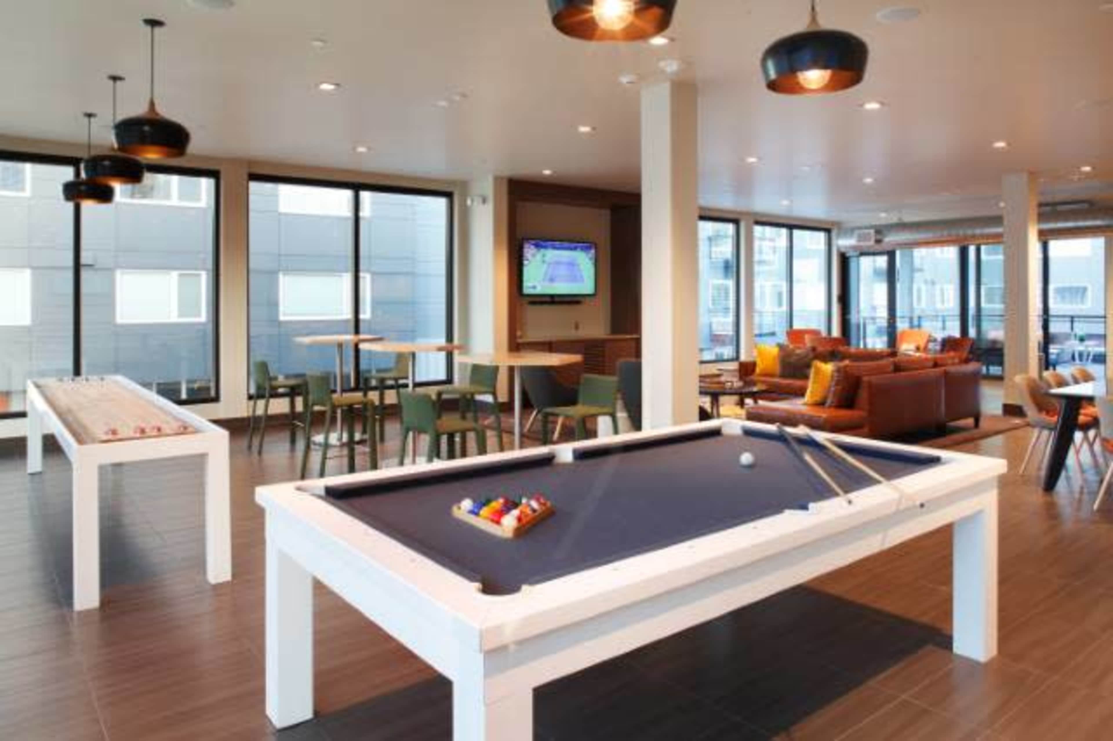 A modern recreational room features billiard tables, seating areas, and large windows showing an outdoor view.