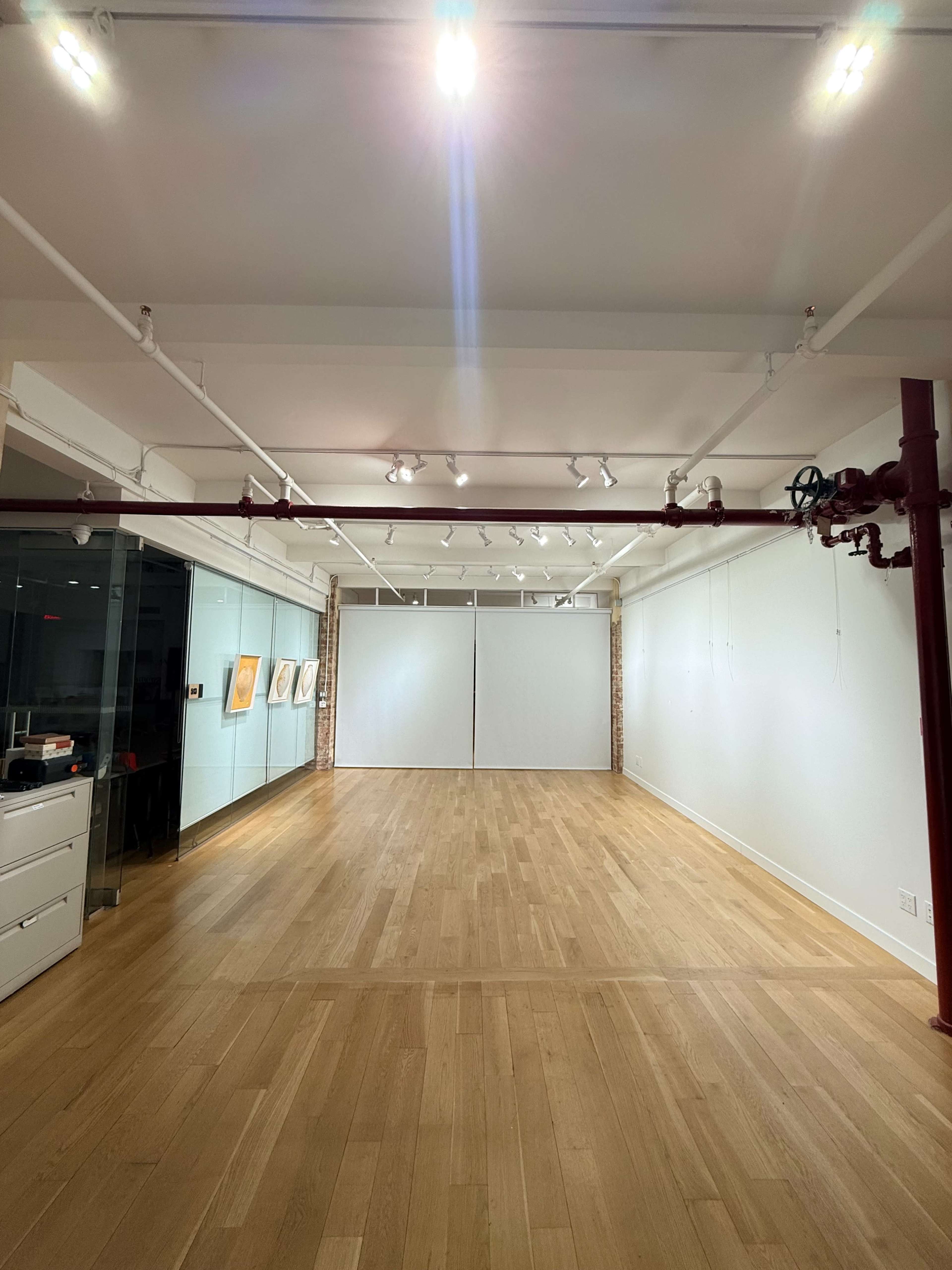 The image shows a spacious, well-lit gallery or studio with wooden flooring and white walls, featuring a large blank section for displays.