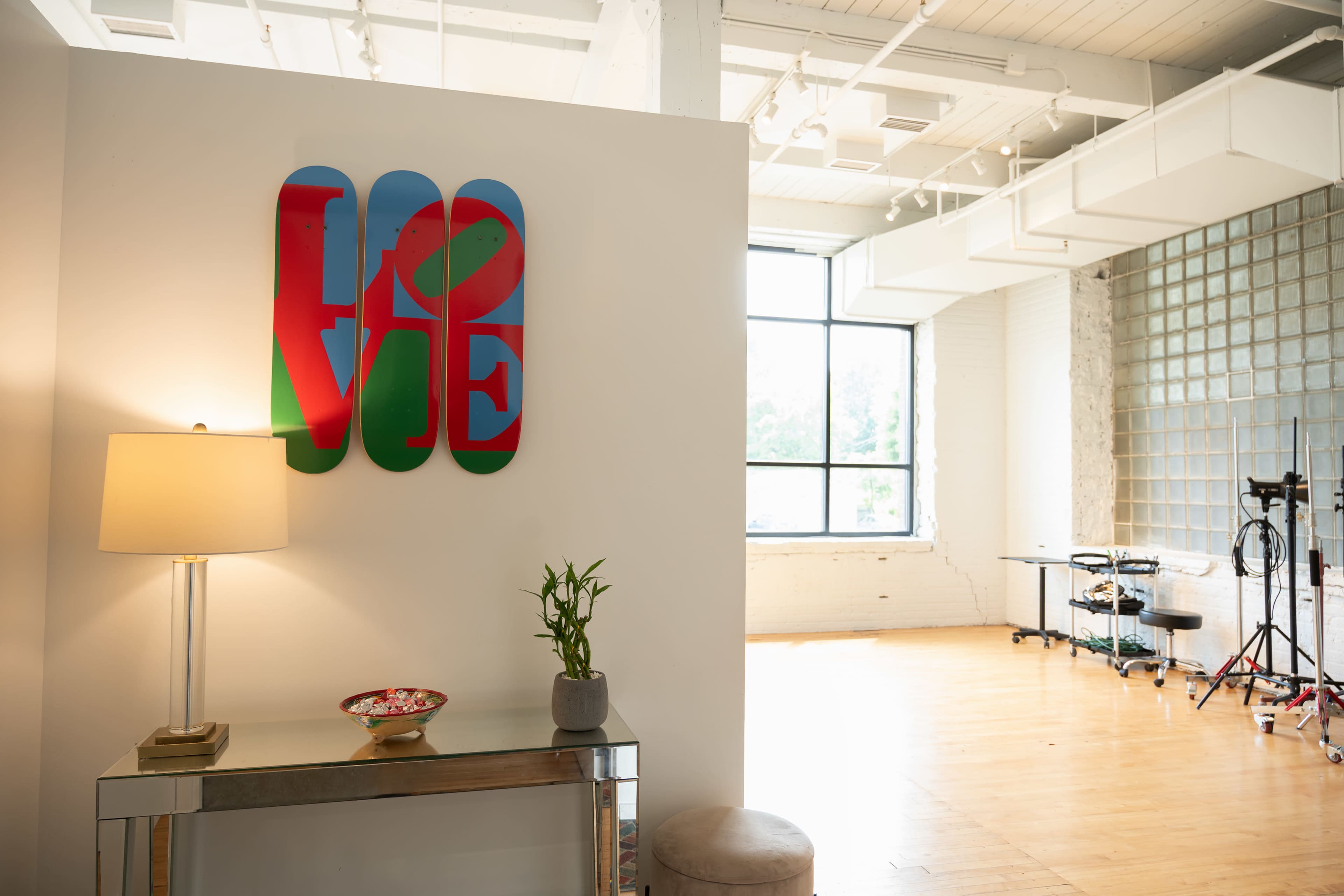 The image shows a modern interior space featuring a lamp, a decorative plant, and a colorful "LOVE" wall art near a large window with natural light.