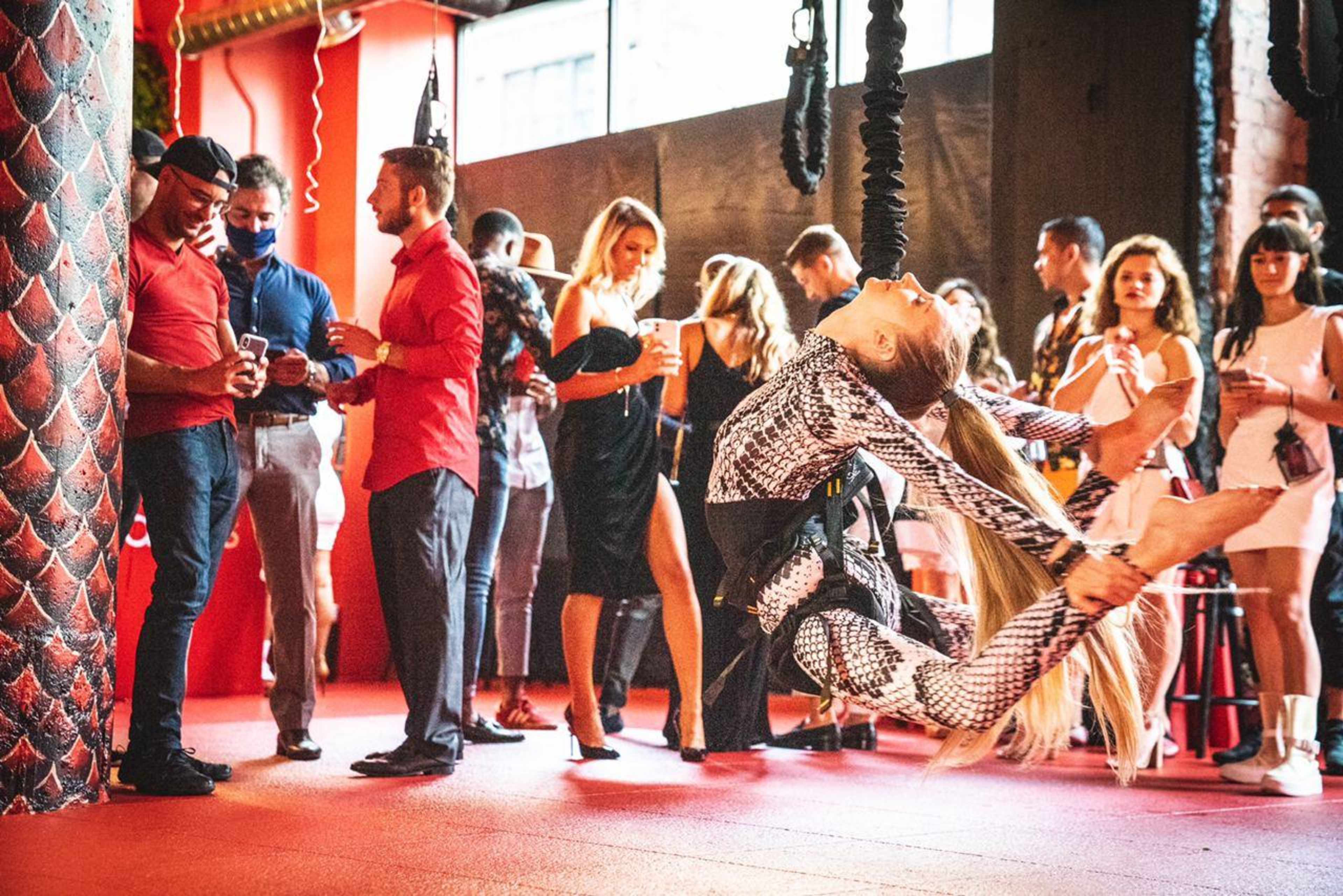 A dancer performs on aerial silks in a lively venue filled with guests mingling and enjoying drinks.