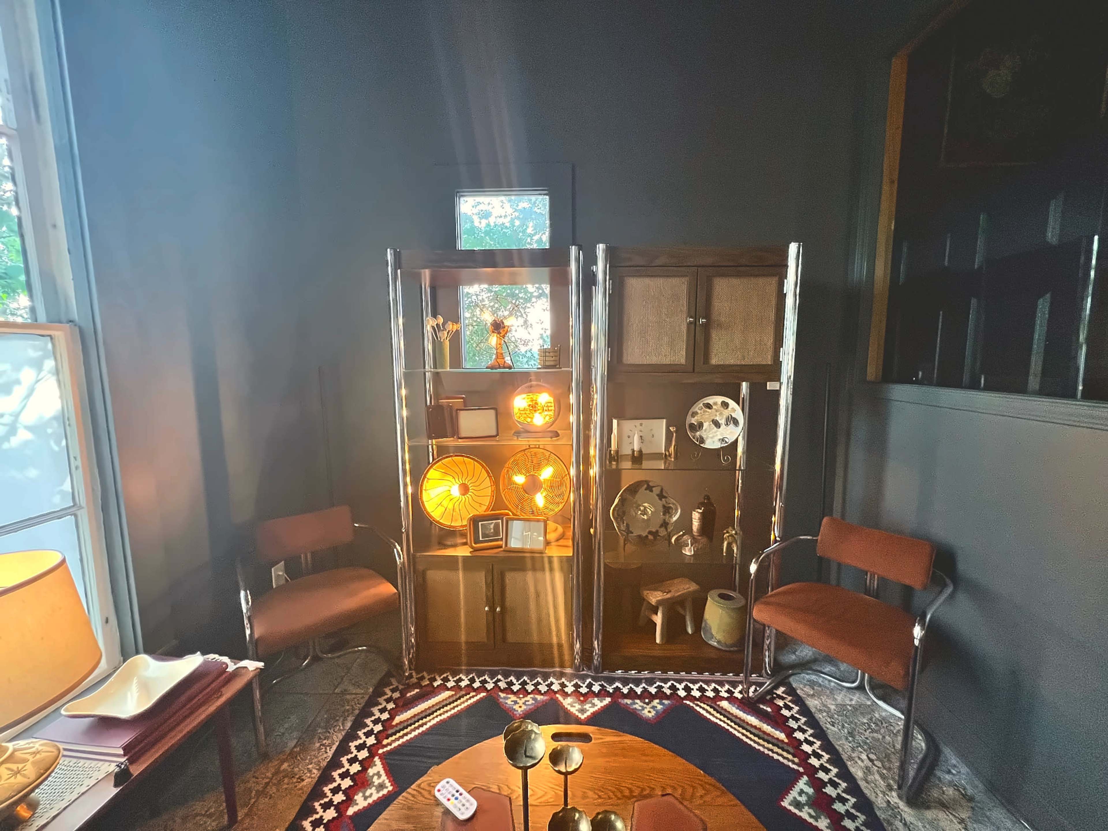 The image shows a cozy room with a display cabinet illuminated by warm lighting, surrounded by two armchairs and a round coffee table.