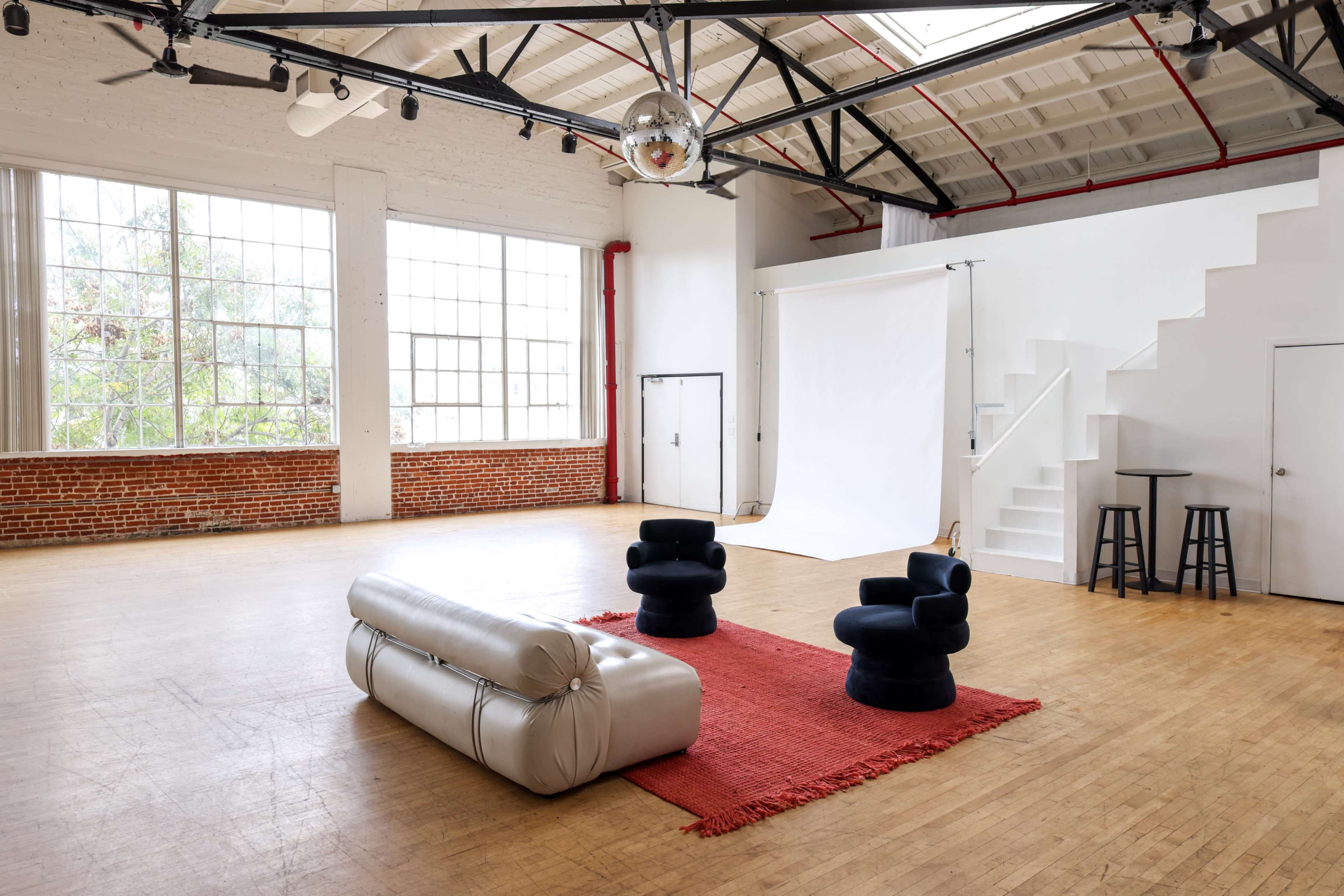 The image shows a spacious, well-lit studio interior featuring a white backdrop, two black upholstered chairs, a silver couch, and a red area rug on the wooden floor.