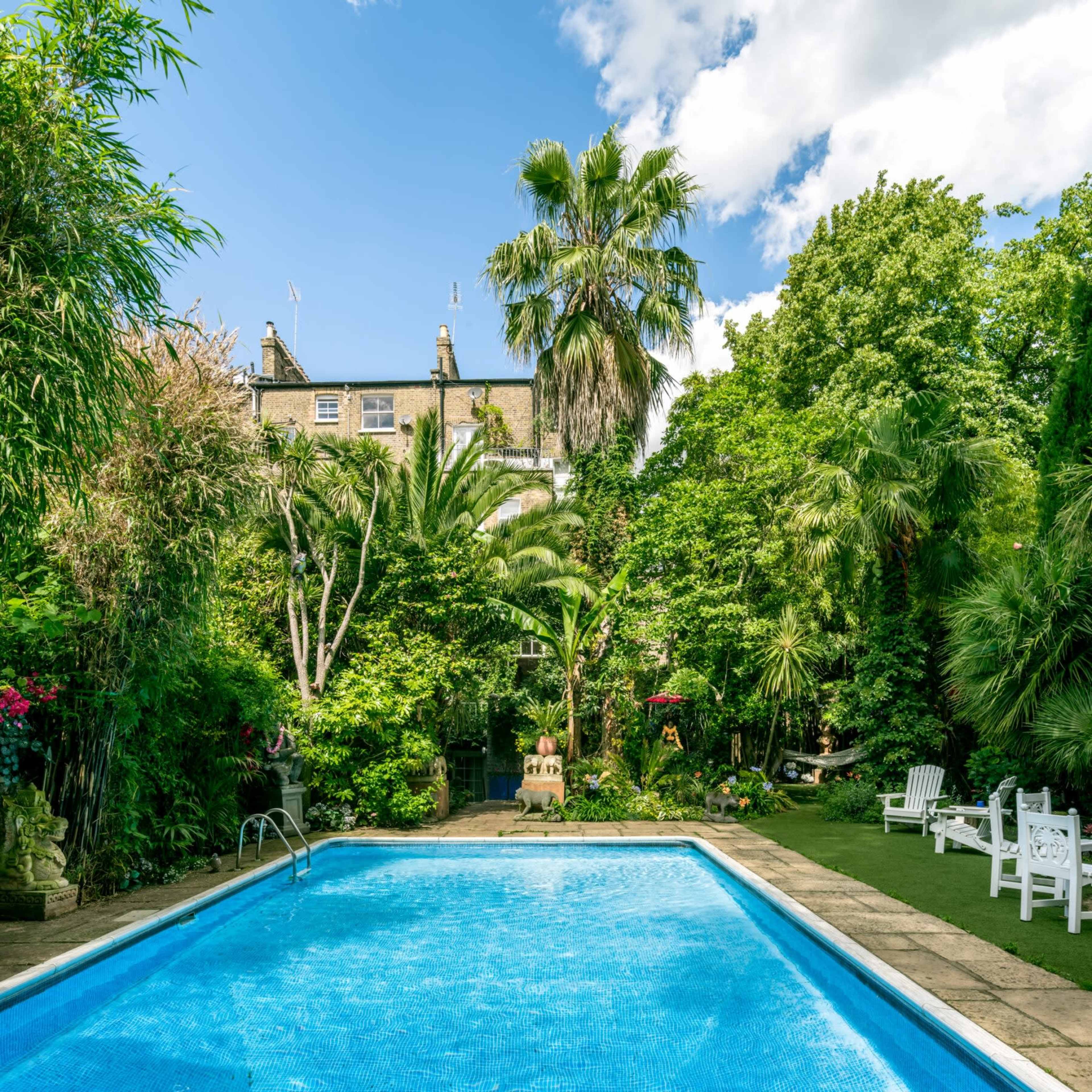 The image shows a swimming pool surrounded by lush greenery and palm trees, with a building partially visible in the background.