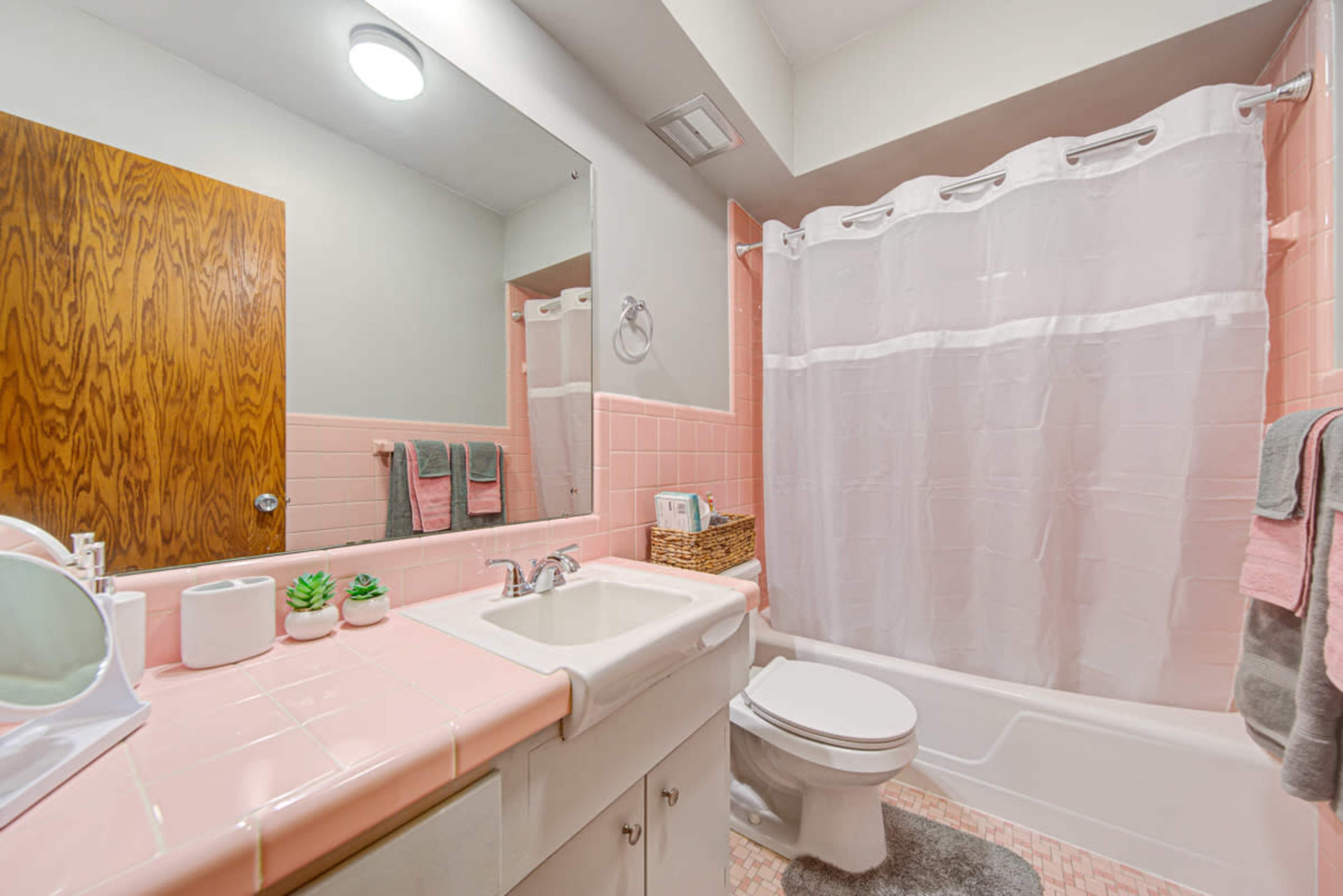 The image shows a small bathroom featuring pink tile accents, a white shower curtain, and basic fixtures including a sink and a toilet.