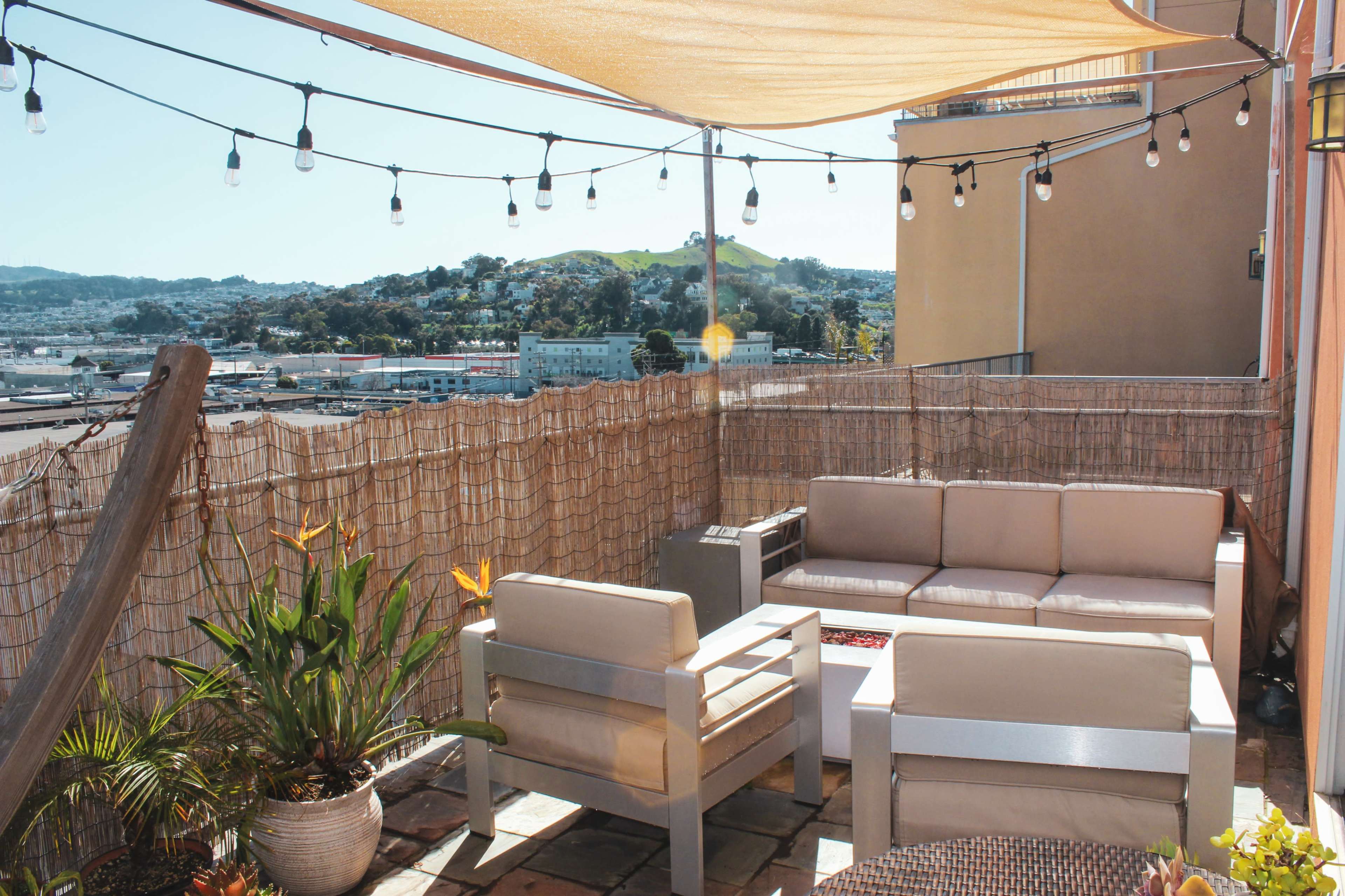 A rooftop patio features a seating area with metallic furniture, decorative plants, and string lights, surrounded by a view of hills and buildings.