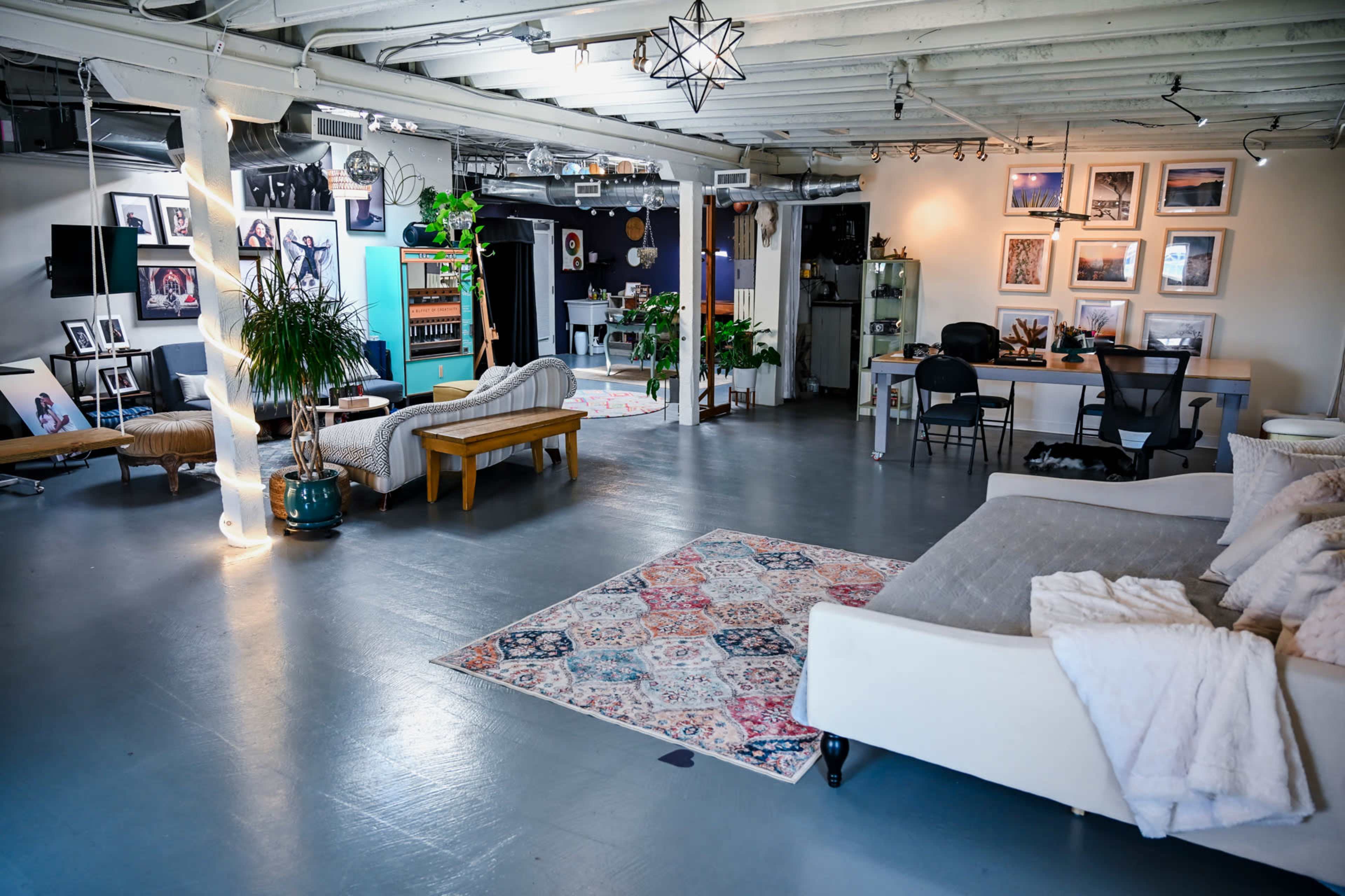 The image shows a spacious, well-lit studio or office area with a mix of seating arrangements, plants, artwork on the walls, and a desk workspace.