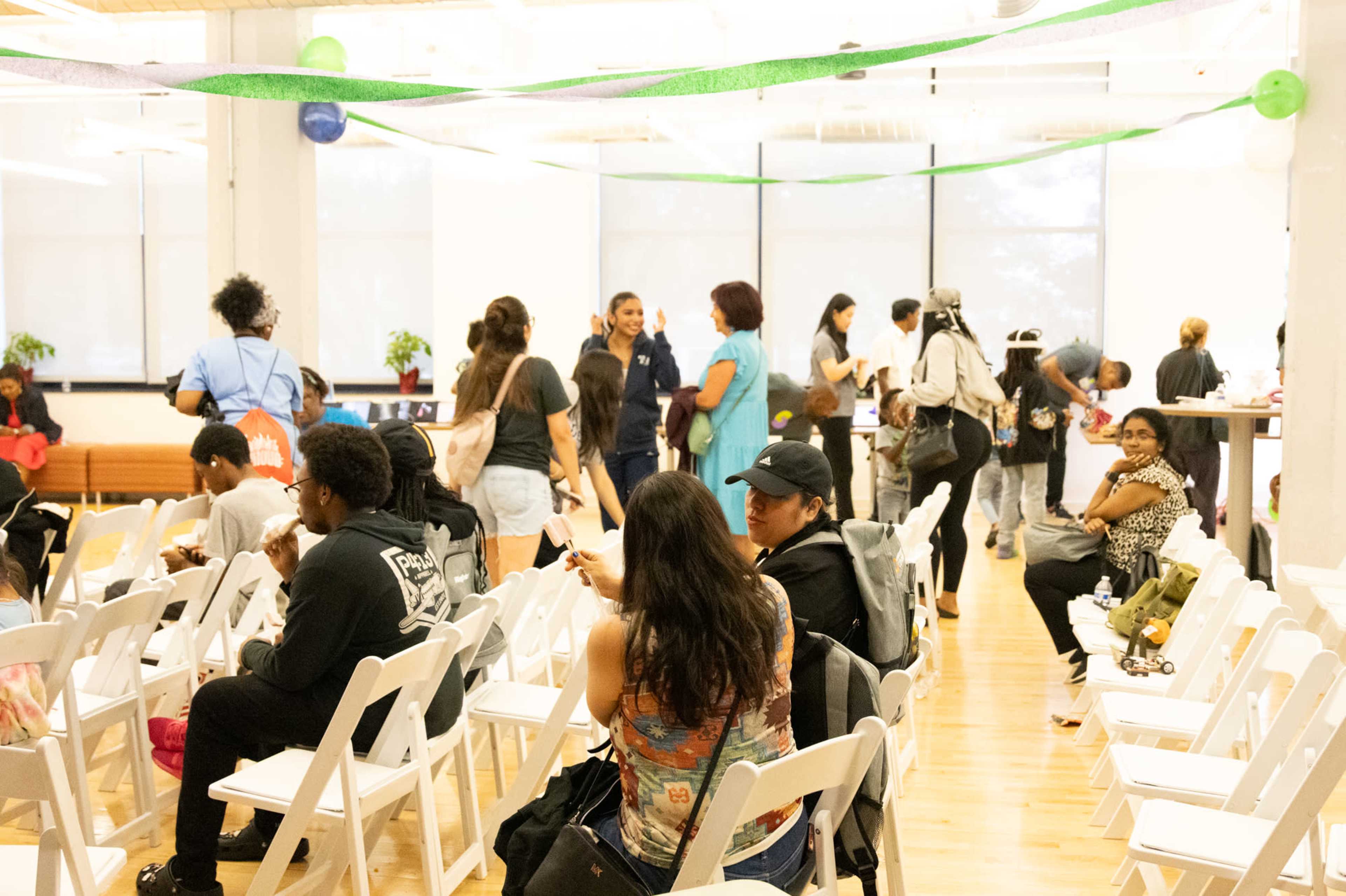 The image shows a spacious indoor event area with several people seated and mingling among white folding chairs, while green decorations hang from the ceiling.
