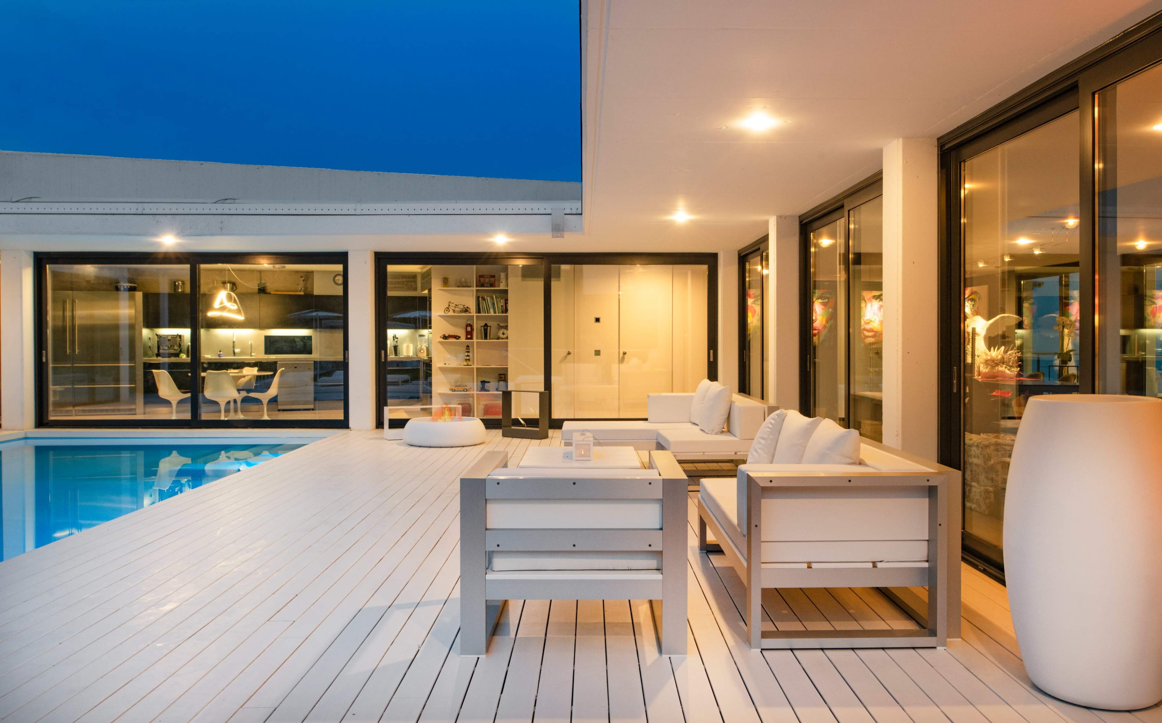 A modern outdoor patio area with seating, overlooking a swimming pool and a well-lit interior space.