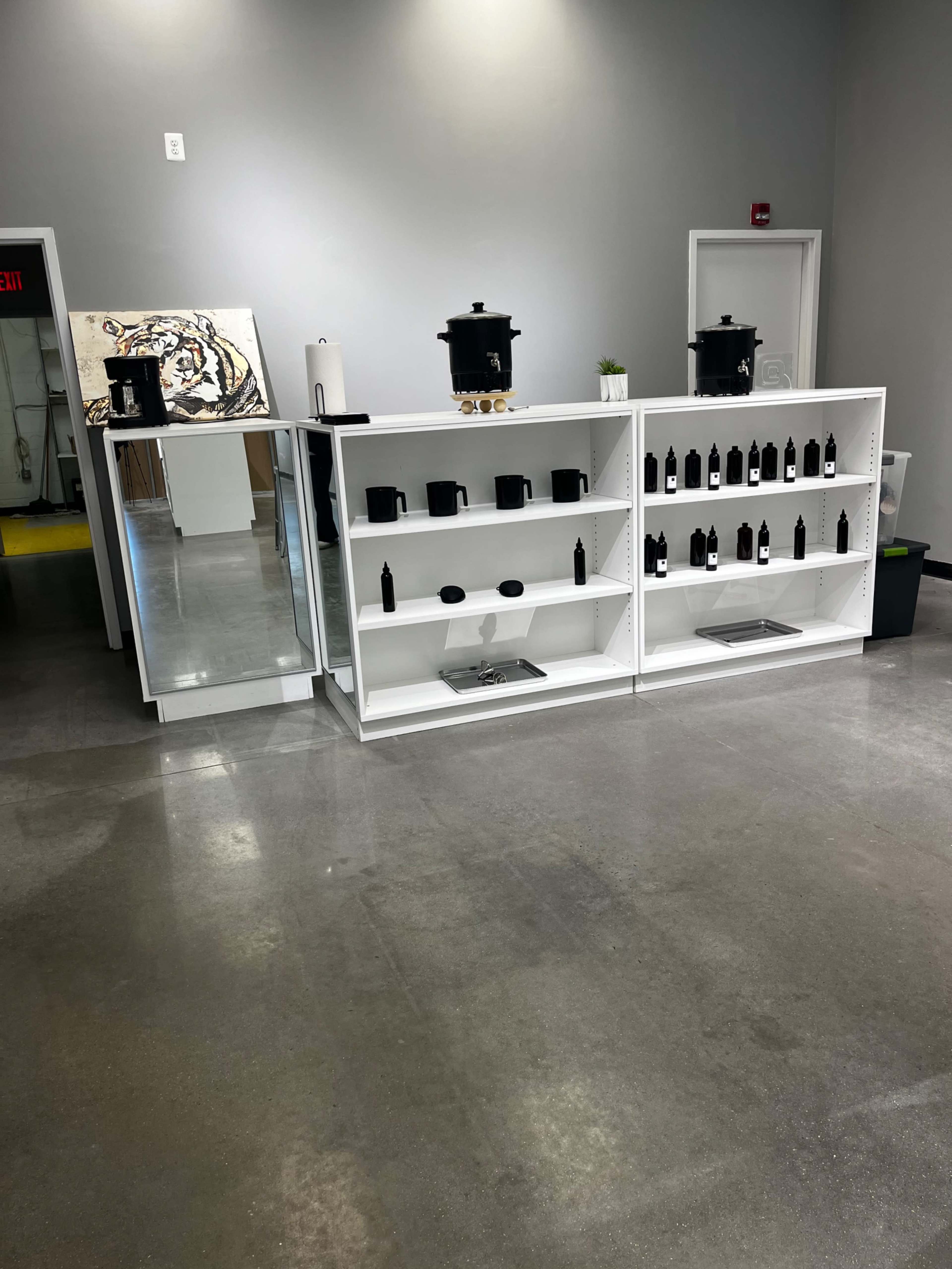 A white shelving unit displays black bottles, with a mirror and two large containers visible in the background of a modern, minimalist room.