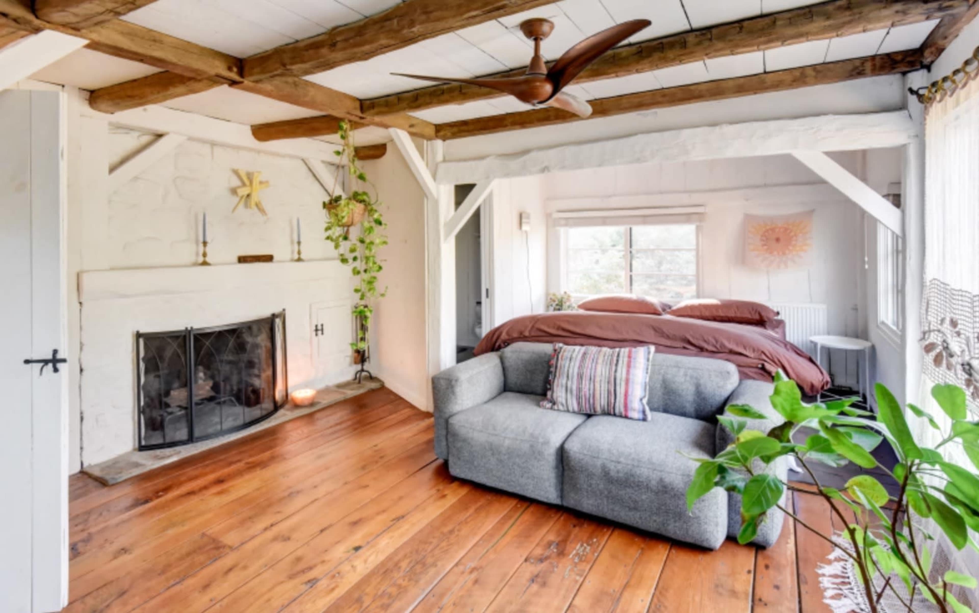 A cozy room with a fireplace, a bed, a gray sofa, large windows, and wooden beams on the ceiling.