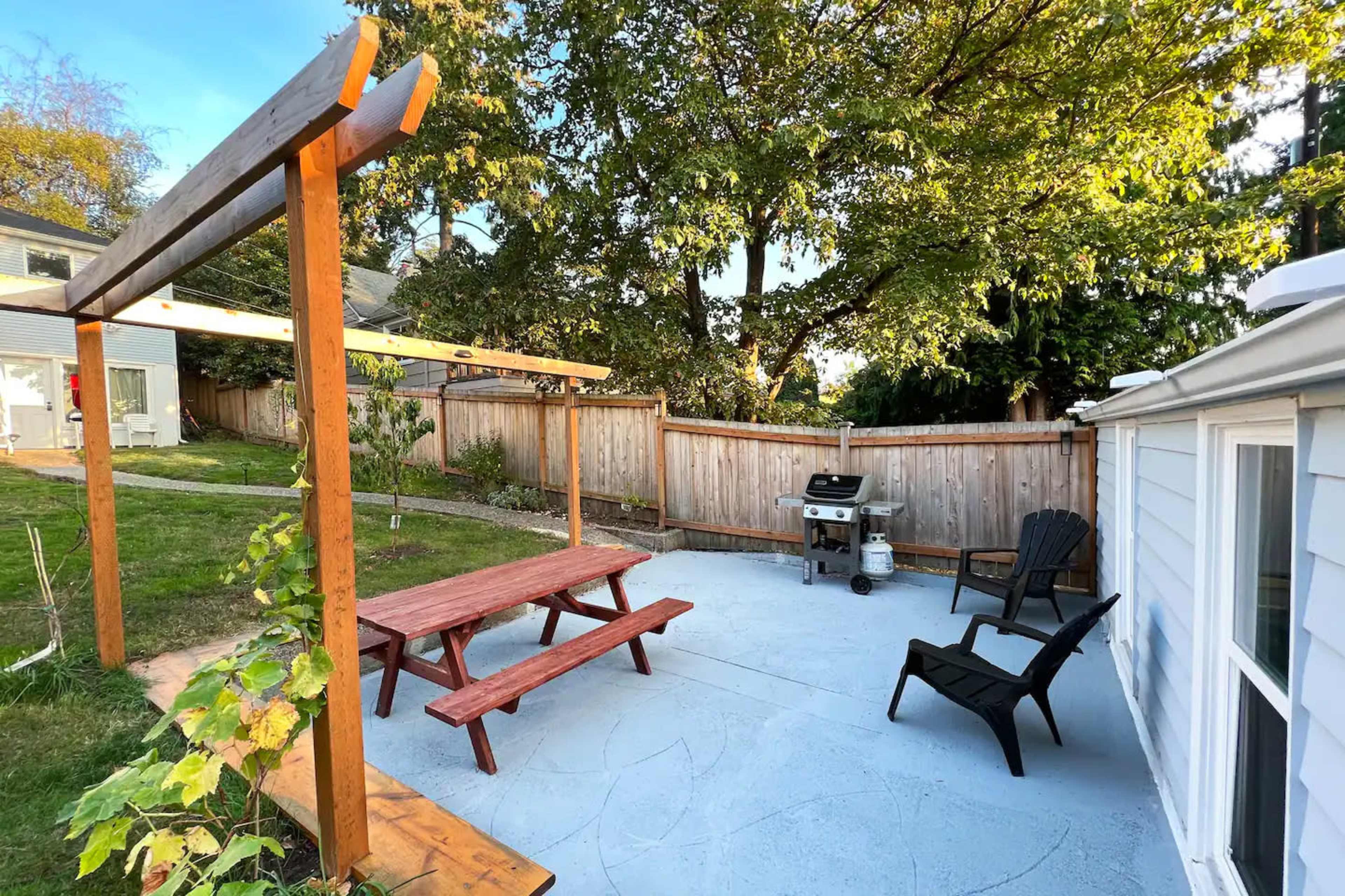 A backyard patio with a picnic table, a grill, and black Adirondack chairs surrounded by trees and a fence.