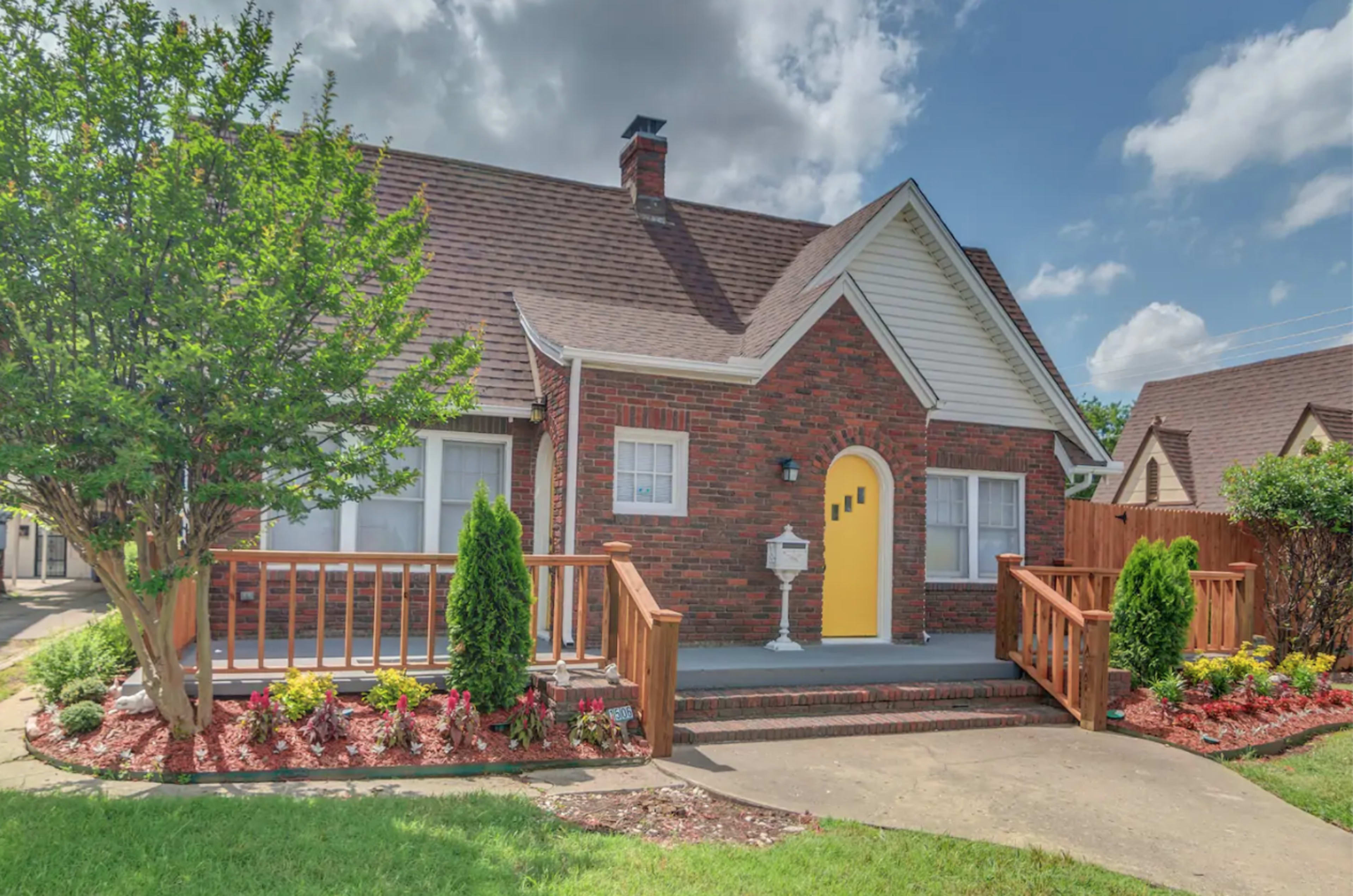 A brick house with a yellow front door and white trim, surrounded by landscaped gardens and a wooden railing leading to the entrance.