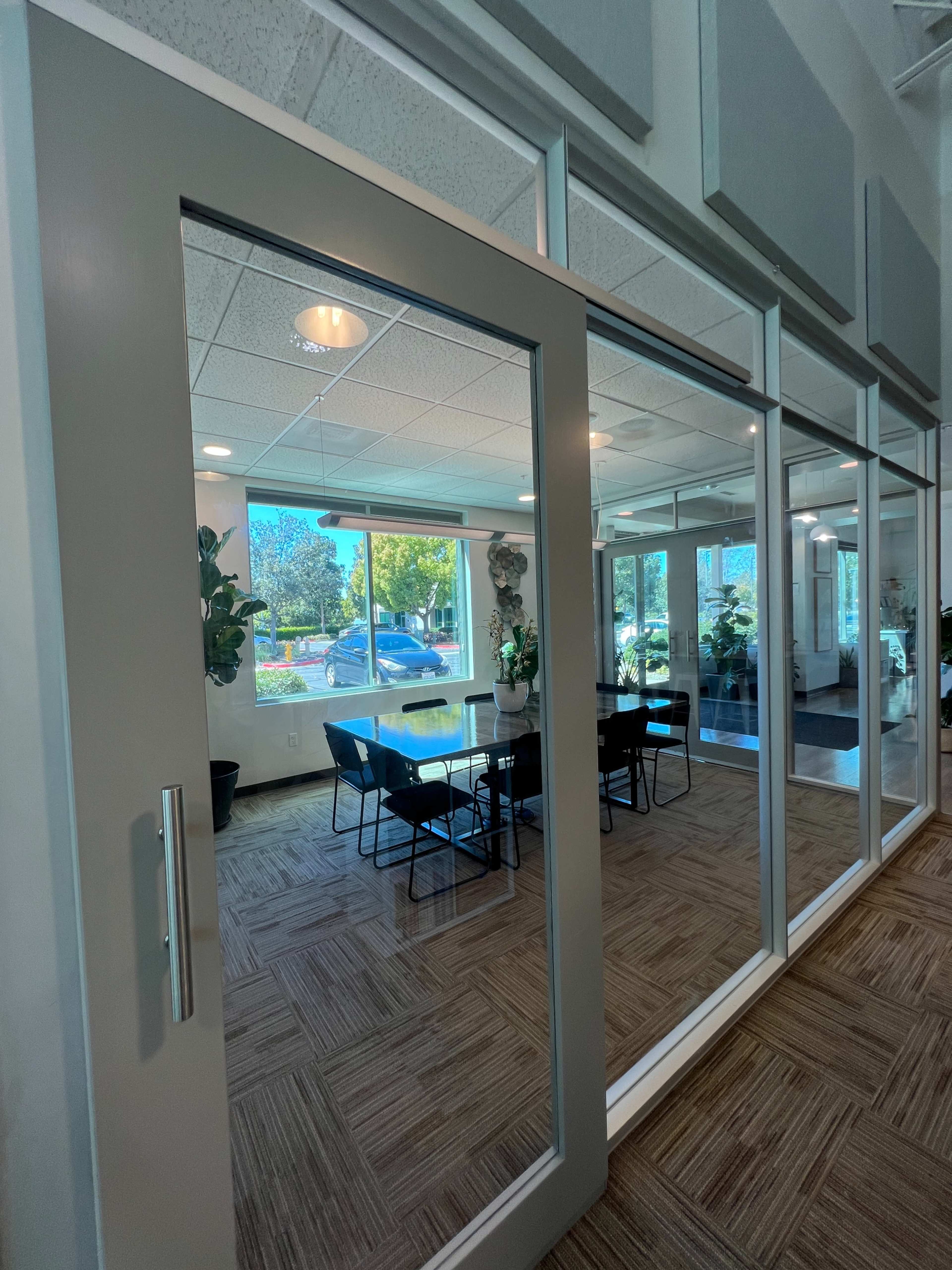 A conference room with a glass door features a large table surrounded by chairs and is illuminated by natural light from nearby windows.