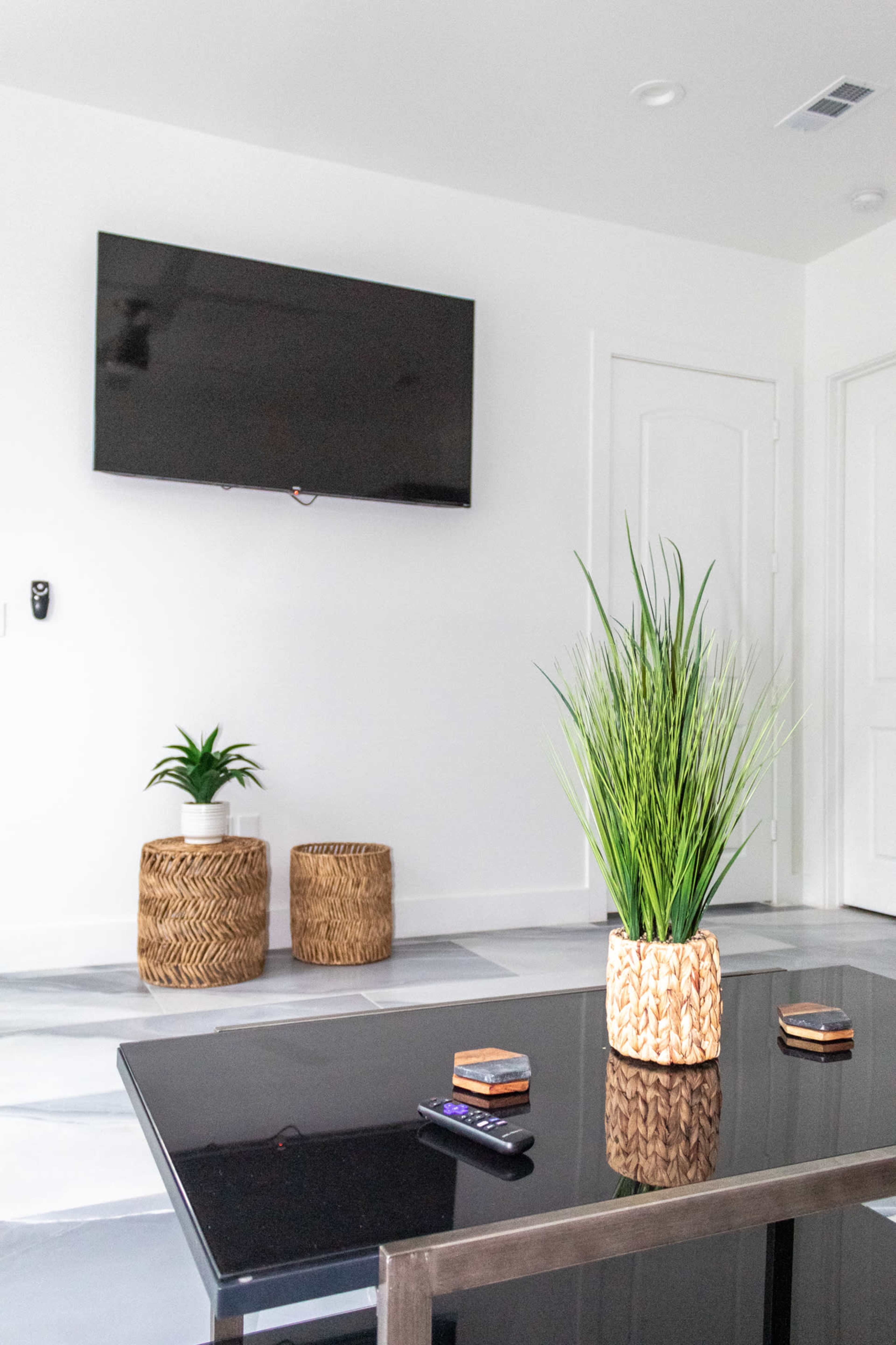 A modern living room features a large flat-screen TV mounted on a white wall, a black table with decorative coasters, and two woven baskets beside potted plants.