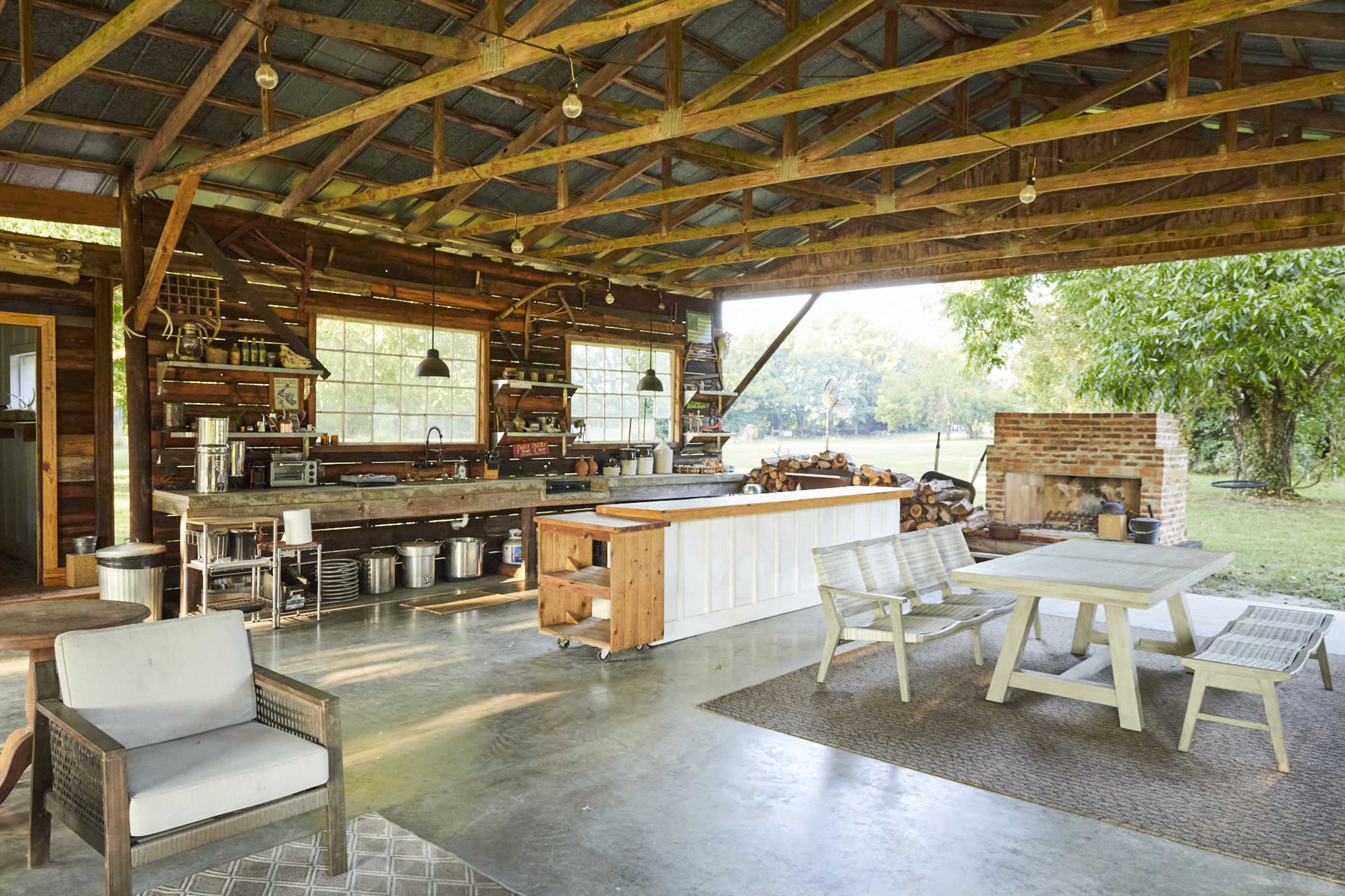 The image shows an open-air rustic kitchen and dining area featuring wooden structures, a bar counter, and a table with chairs under a metal roof.