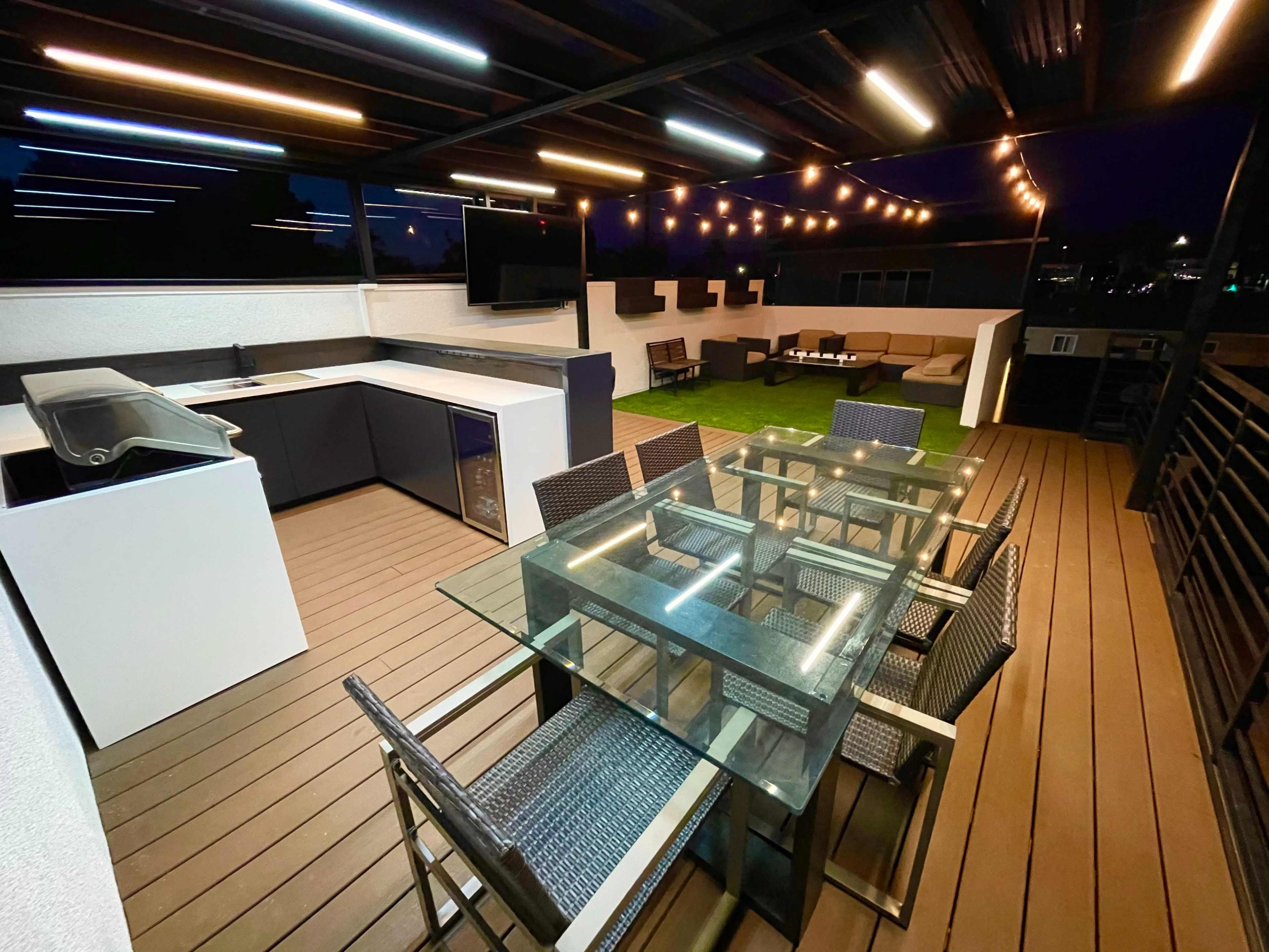 A modern rooftop terrace with a dining table, outdoor kitchen, and lounge area illuminated by string lights at dusk.