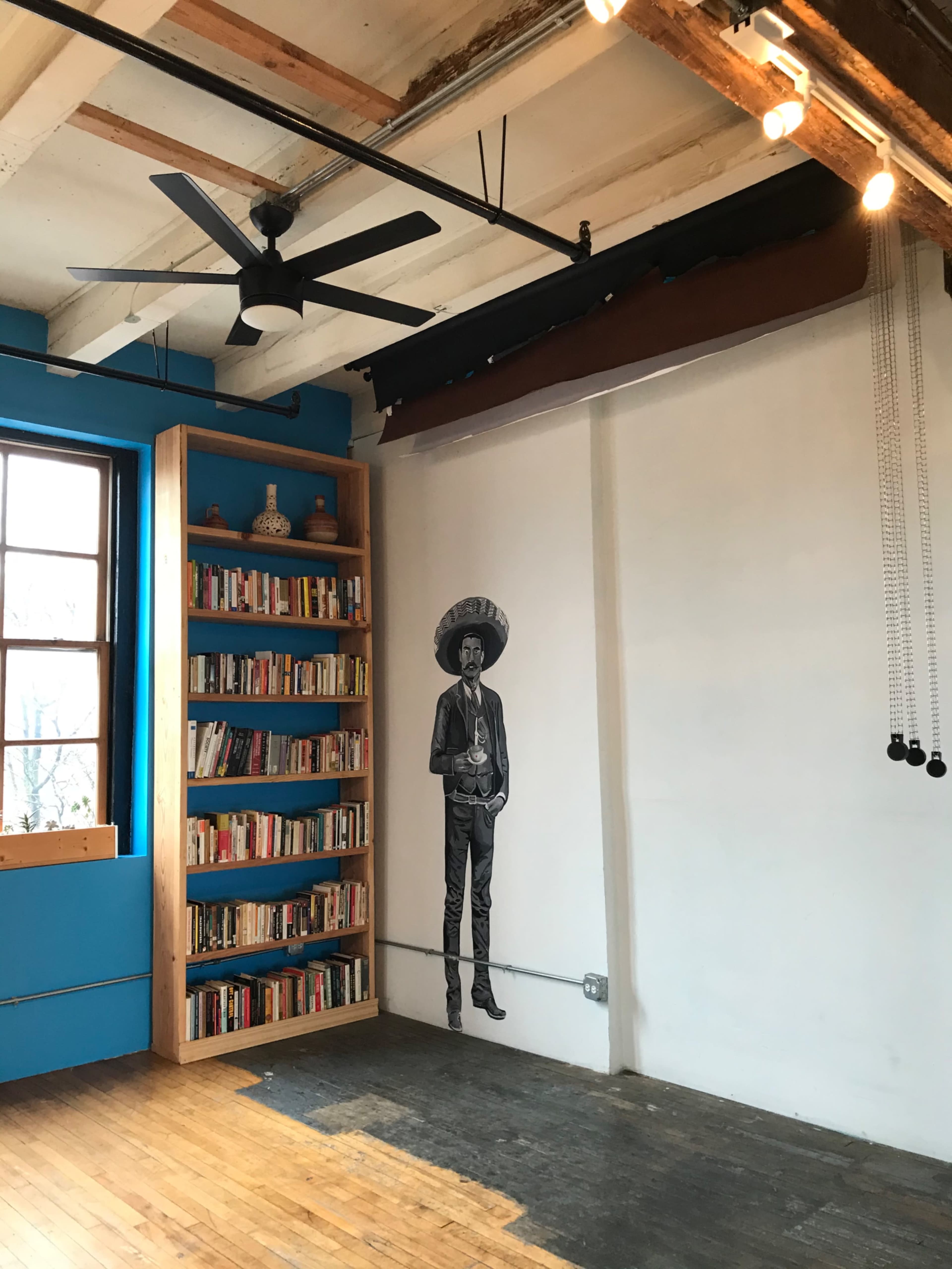 A corner of a room featuring a large wooden bookshelf filled with books and a wall with a mural of a person wearing a sombrero.