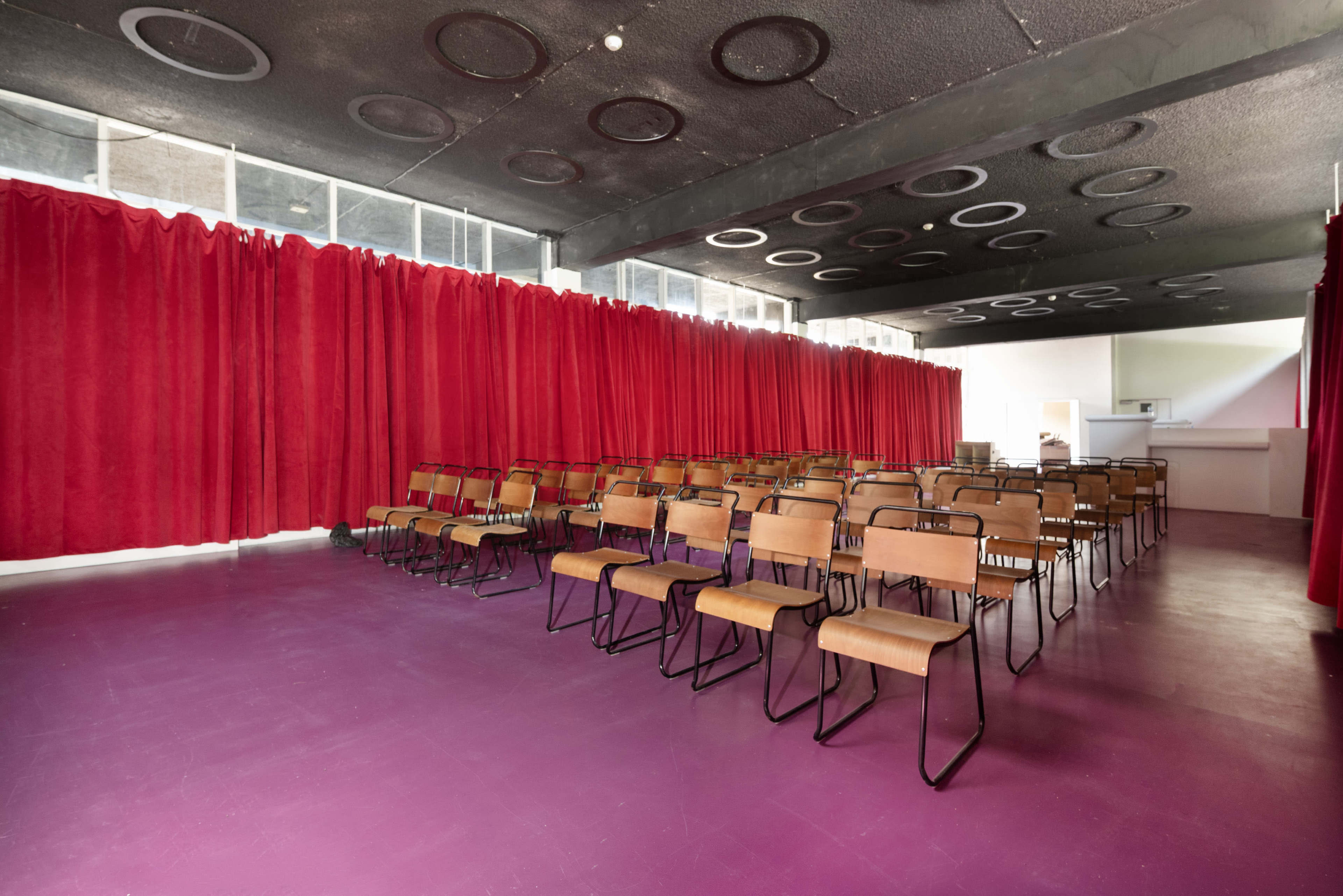 The image shows a spacious room with a pink floor, several rows of brown chairs, and red curtains along the walls.