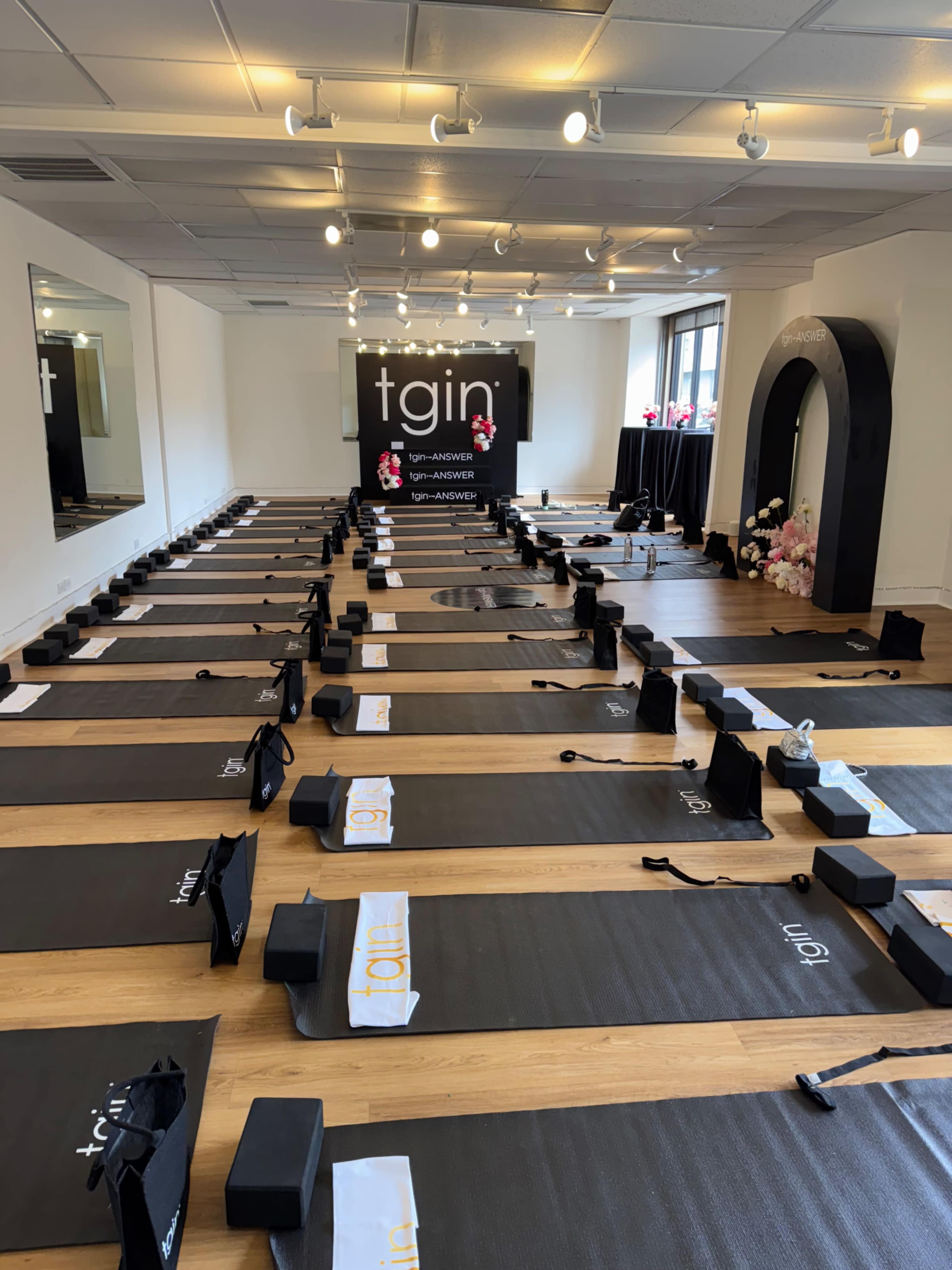 The image shows a yoga studio set up with multiple mats arranged in rows, with a backdrop displaying the brand "tgin" and decorative elements at the front.