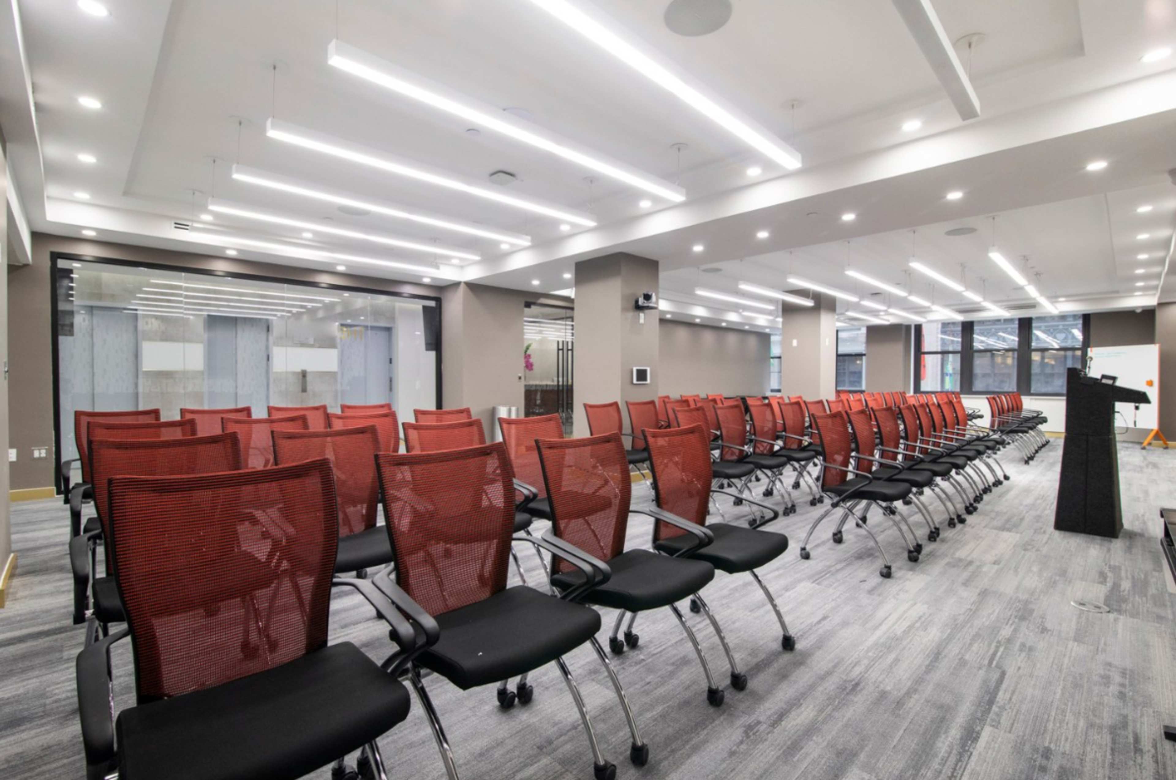 A modern conference room features rows of red mesh office chairs and a black podium under bright overhead lighting.