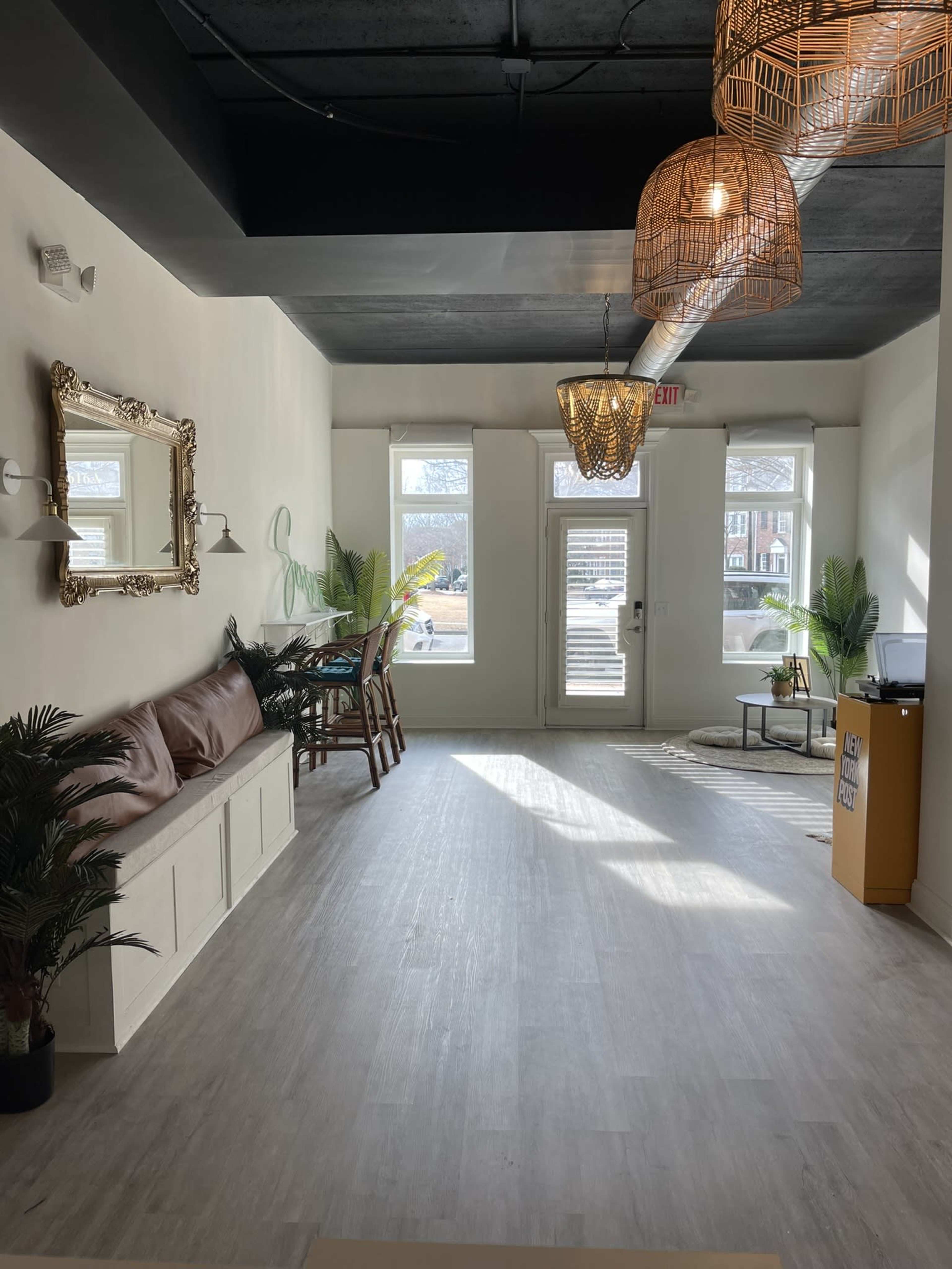 A bright, modern interior of a room featuring a seating area with a brown couch, plants, and large windows letting in natural light.