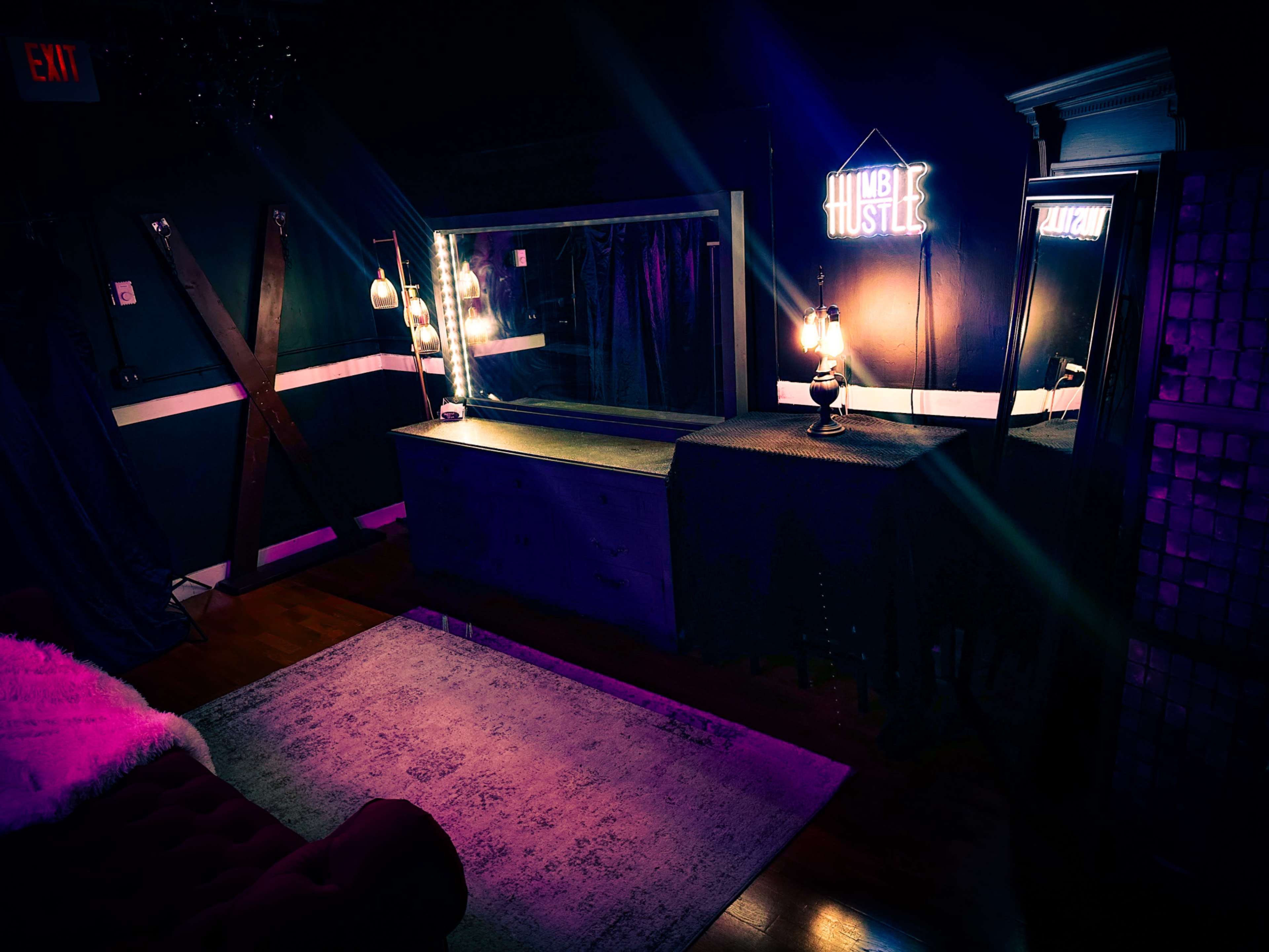 The scene features a dimly lit room with a mirrored vanity, colorful neon signage, and a plush area rug on a wooden floor.