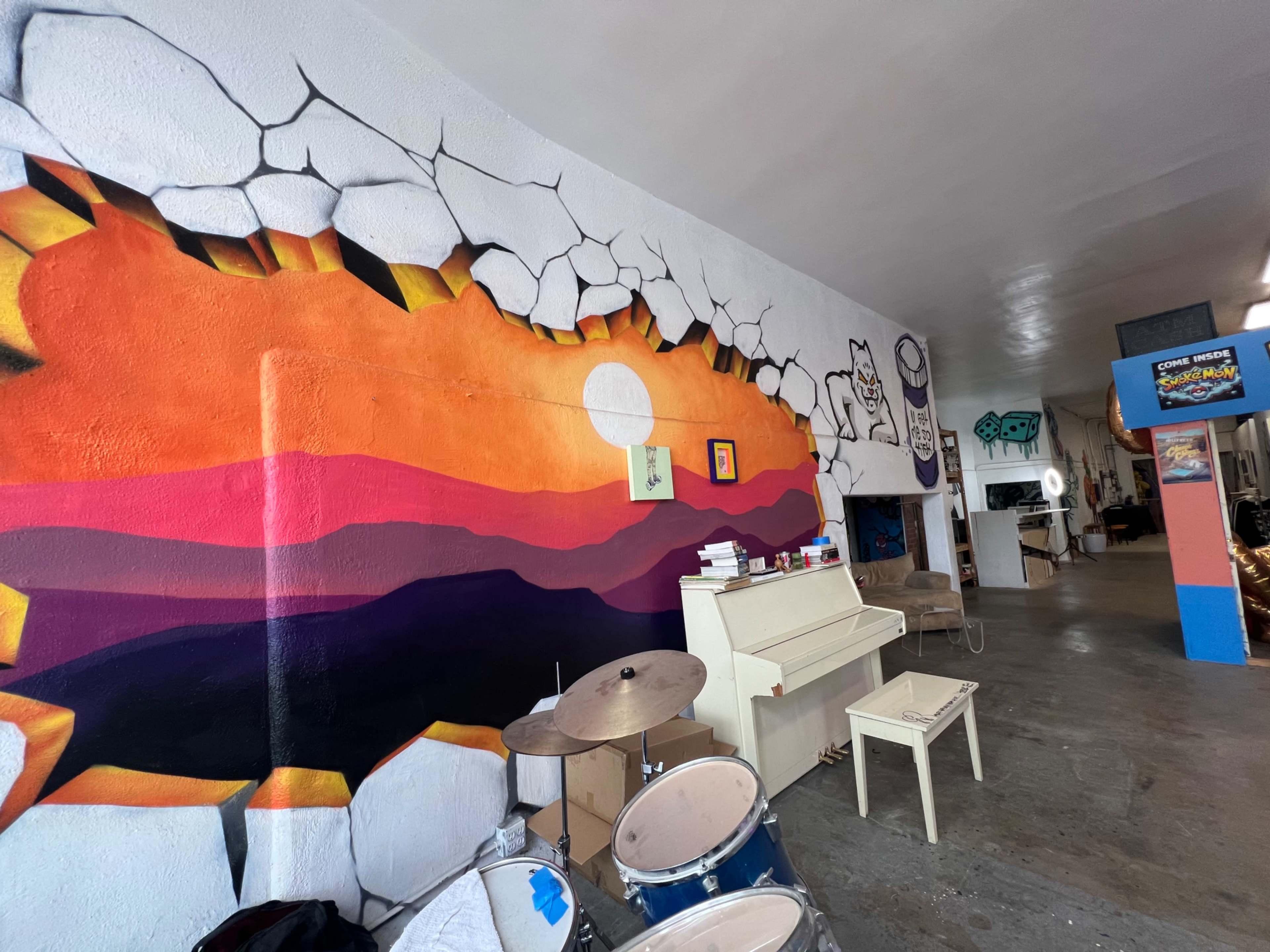 The image shows an indoor space featuring a colorful mural of a sunset, a white piano, and various musical instruments in a creative studio environment.