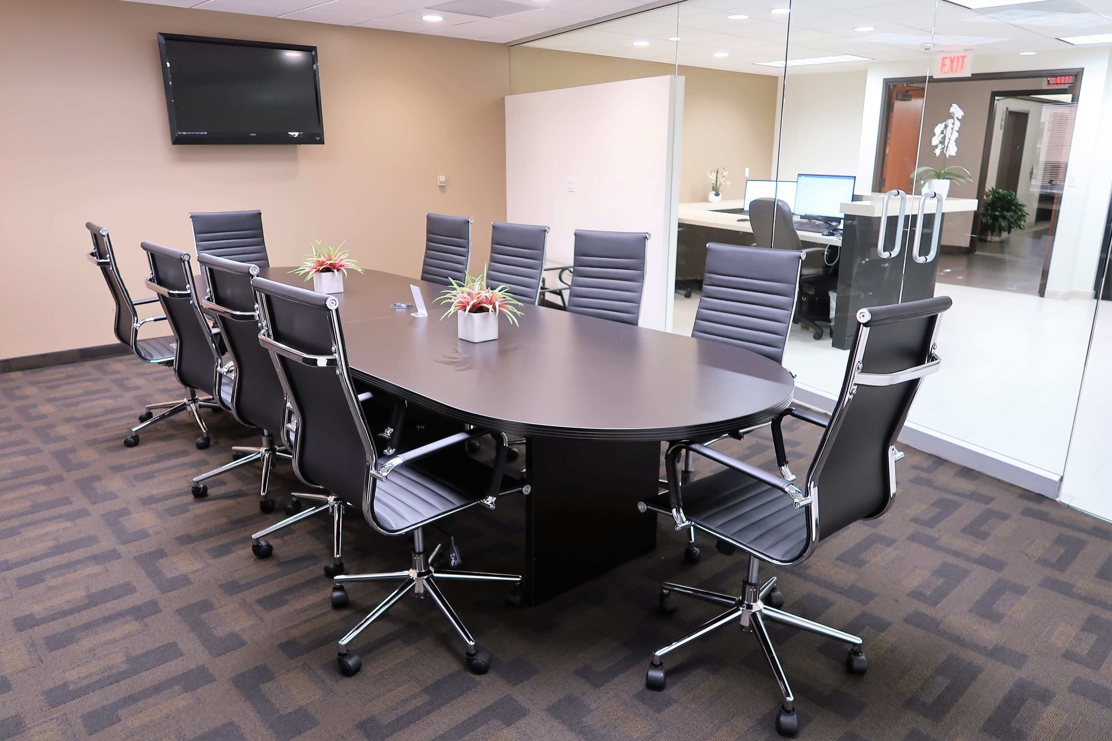 A modern conference room features a large oval table surrounded by sleek black chairs and is separated by a glass wall from an adjacent workspace.