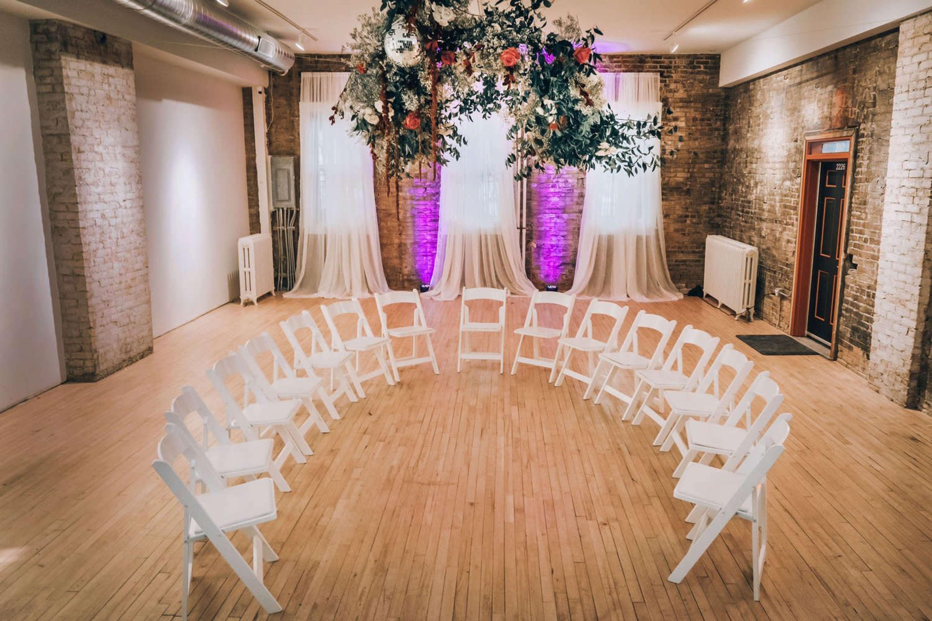 A circular arrangement of white chairs in an indoor space, with draped fabric and floral decorations above.