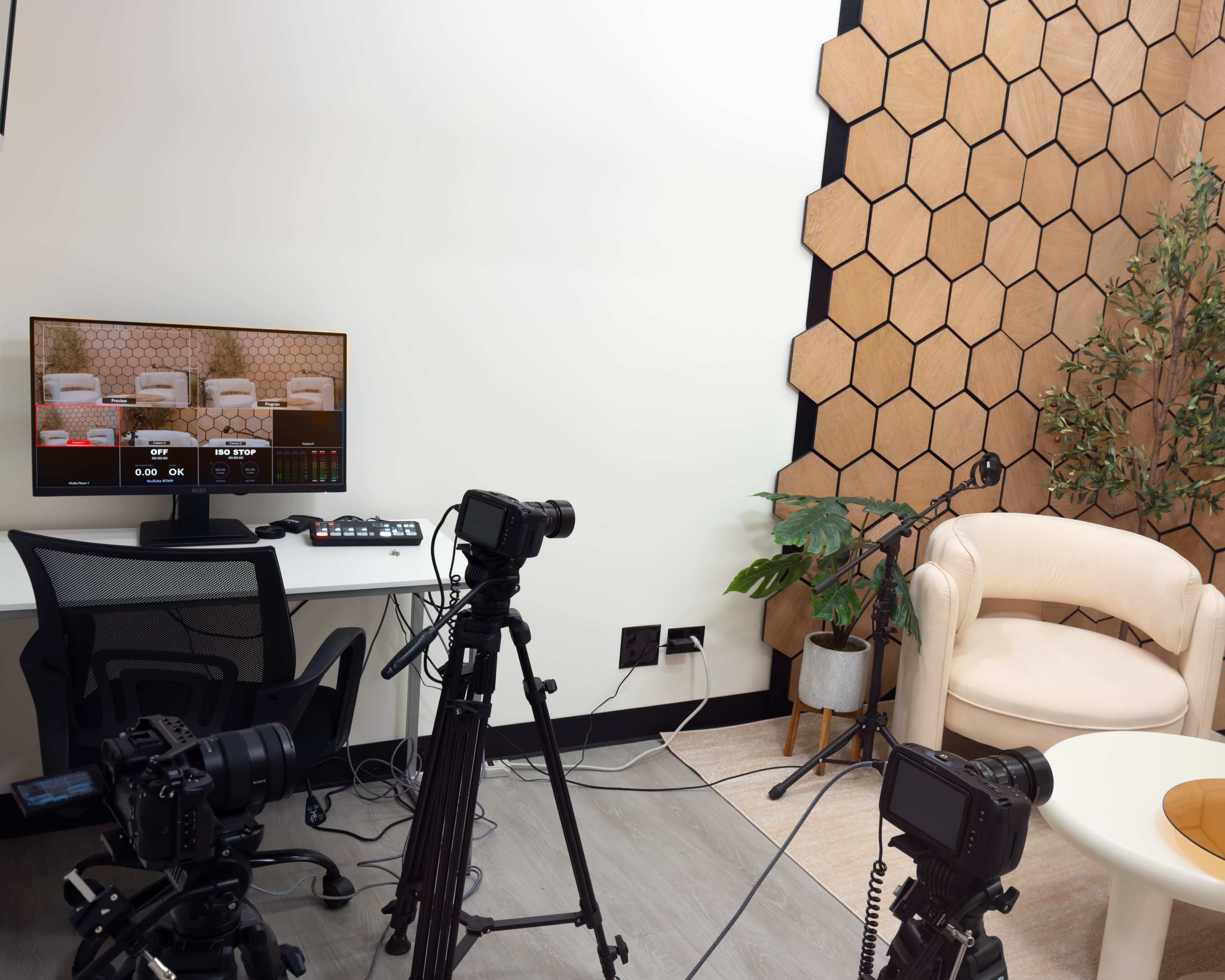 The image shows a recording setup with multiple cameras focused on a stylish, light-colored armchair in a room featuring a hexagonal wall design and a small plant.