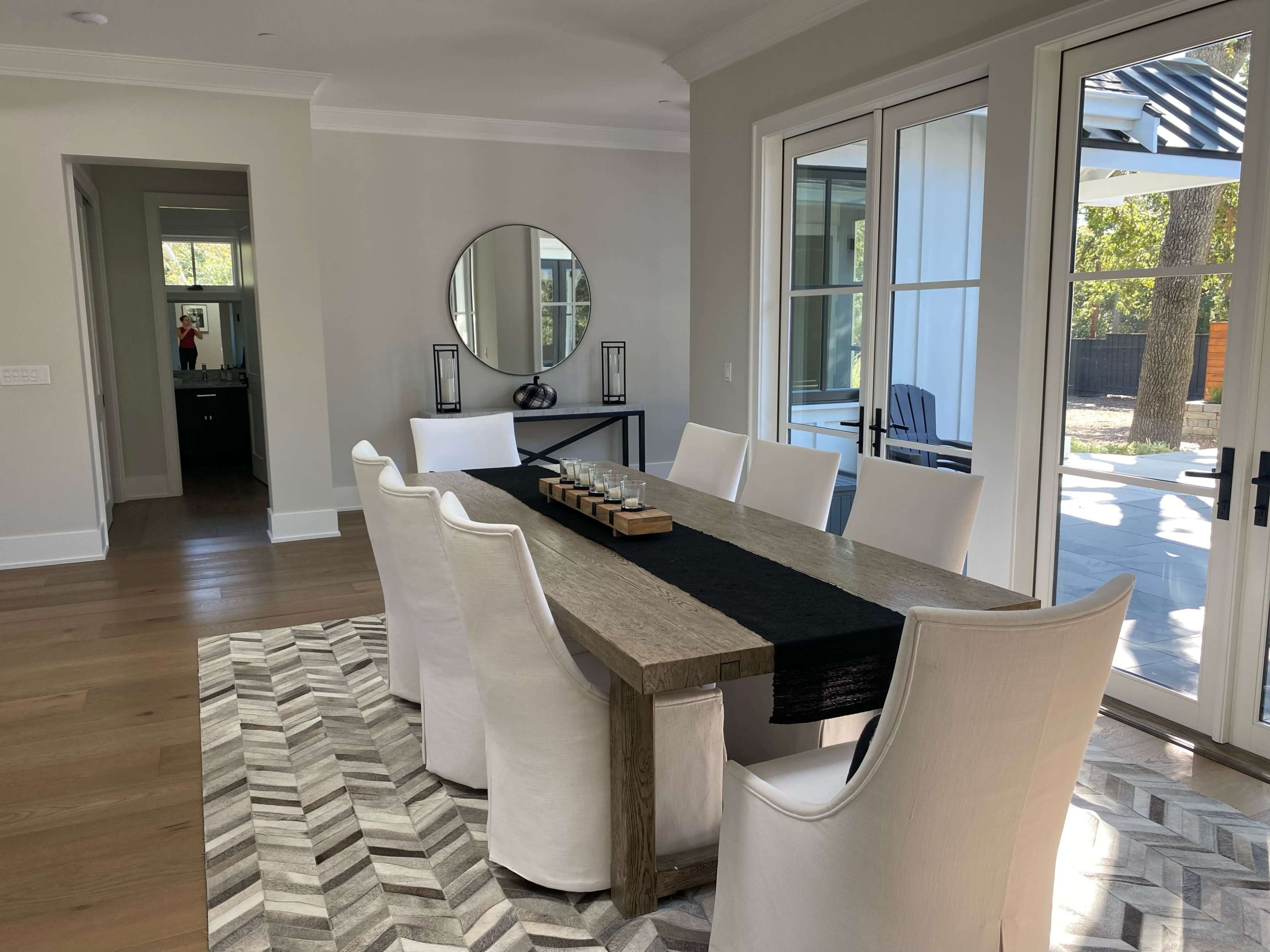 A dining room with a large wooden table surrounded by white chairs, a black runner on the table, and large windows facing an outdoor area.