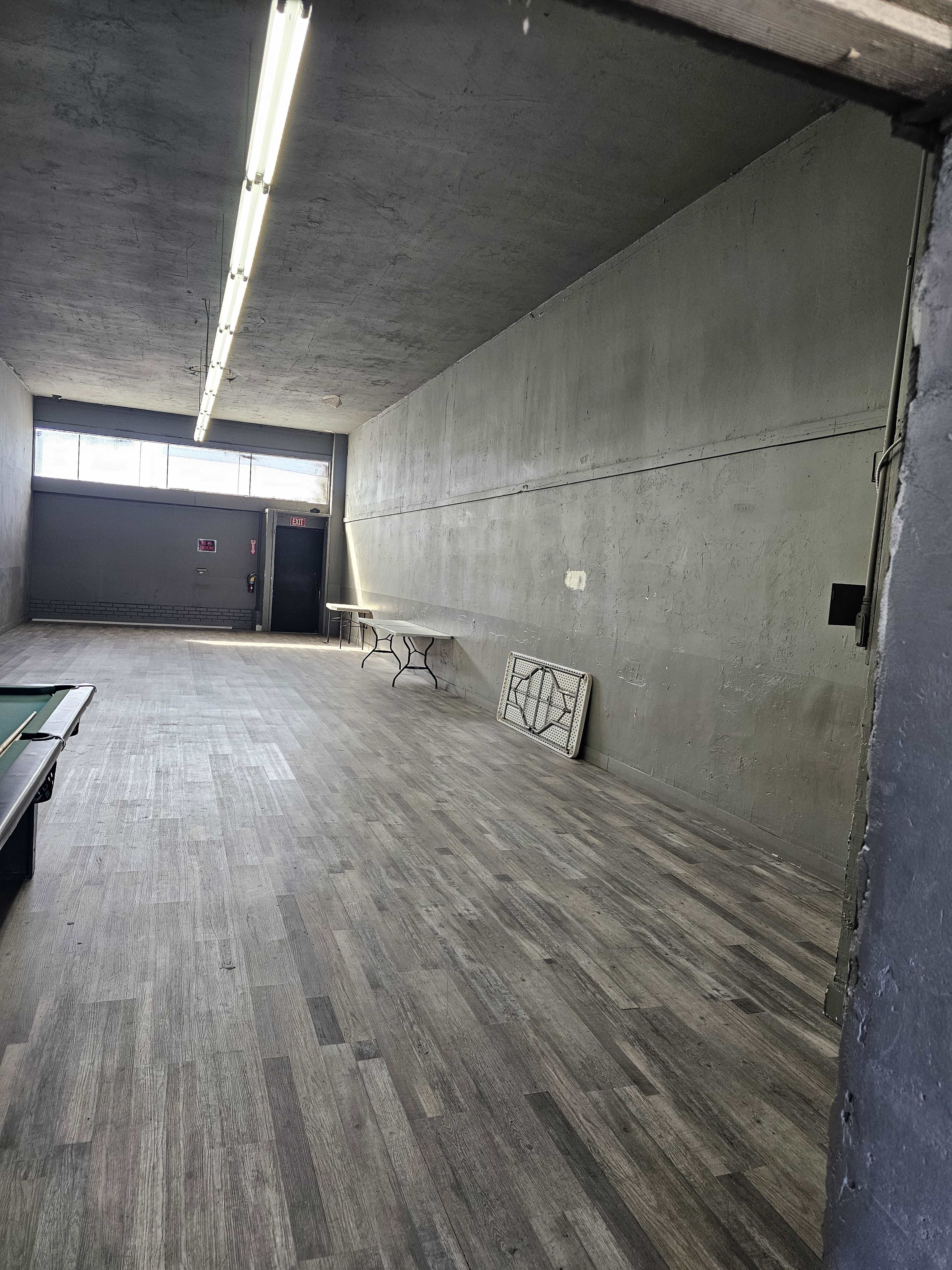 The image shows an empty room with a wooden floor, gray walls, fluorescent lighting, and a table against one wall.