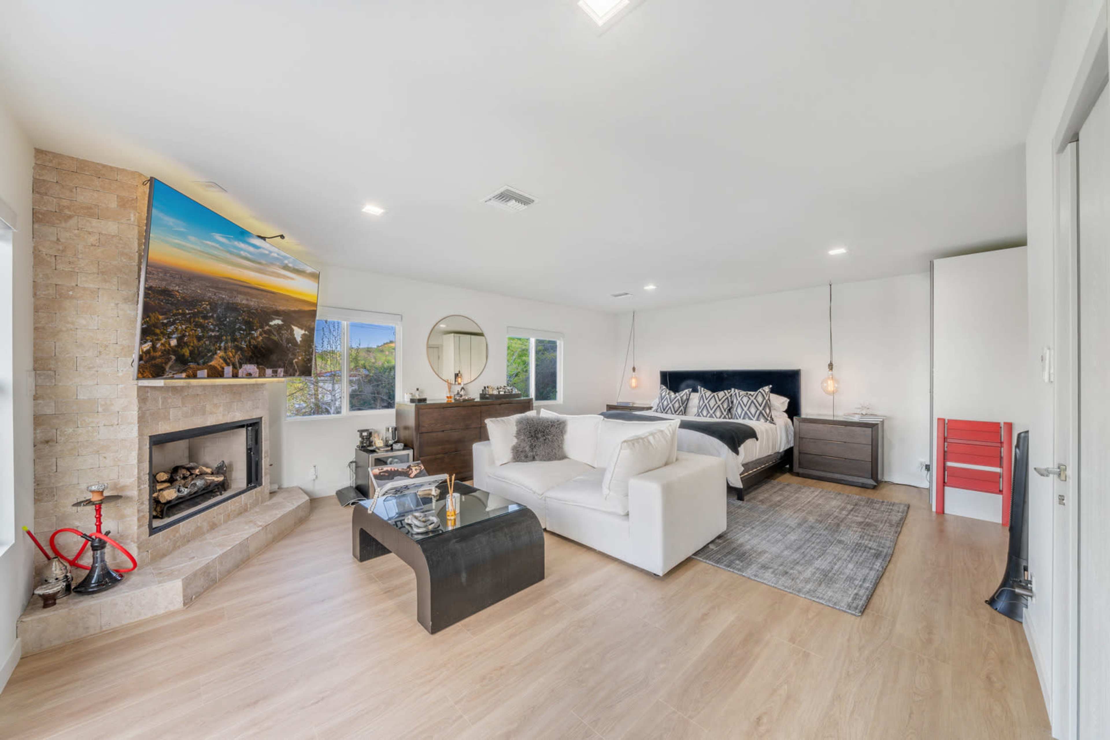 The image shows a modern bedroom with a large bed, a seating area featuring a white couch, a fireplace, and a wall-mounted television displaying a scenic view.