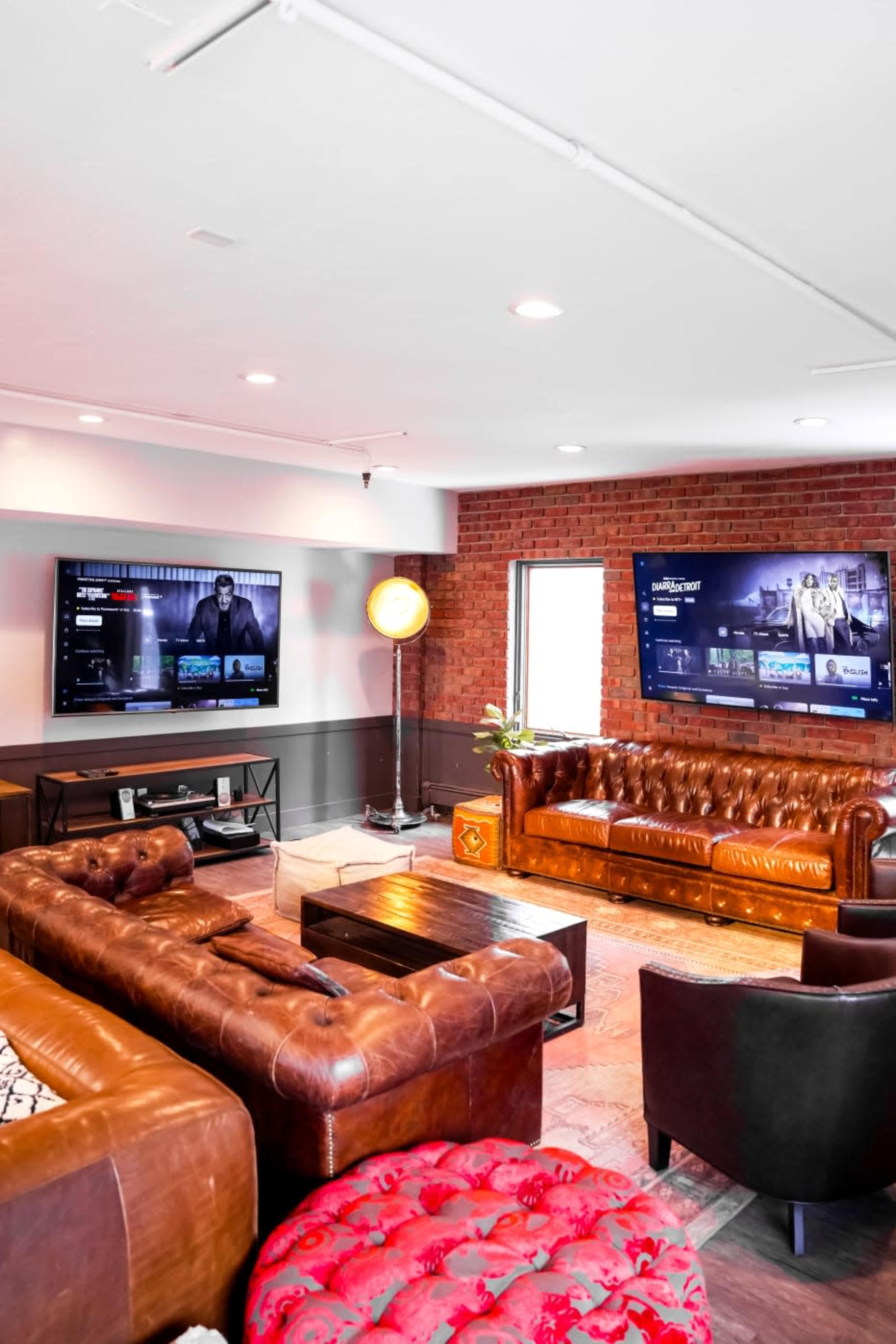 A stylish living room with two large screens mounted on a brick wall, a brown leather sofa, and a red upholstered chair.