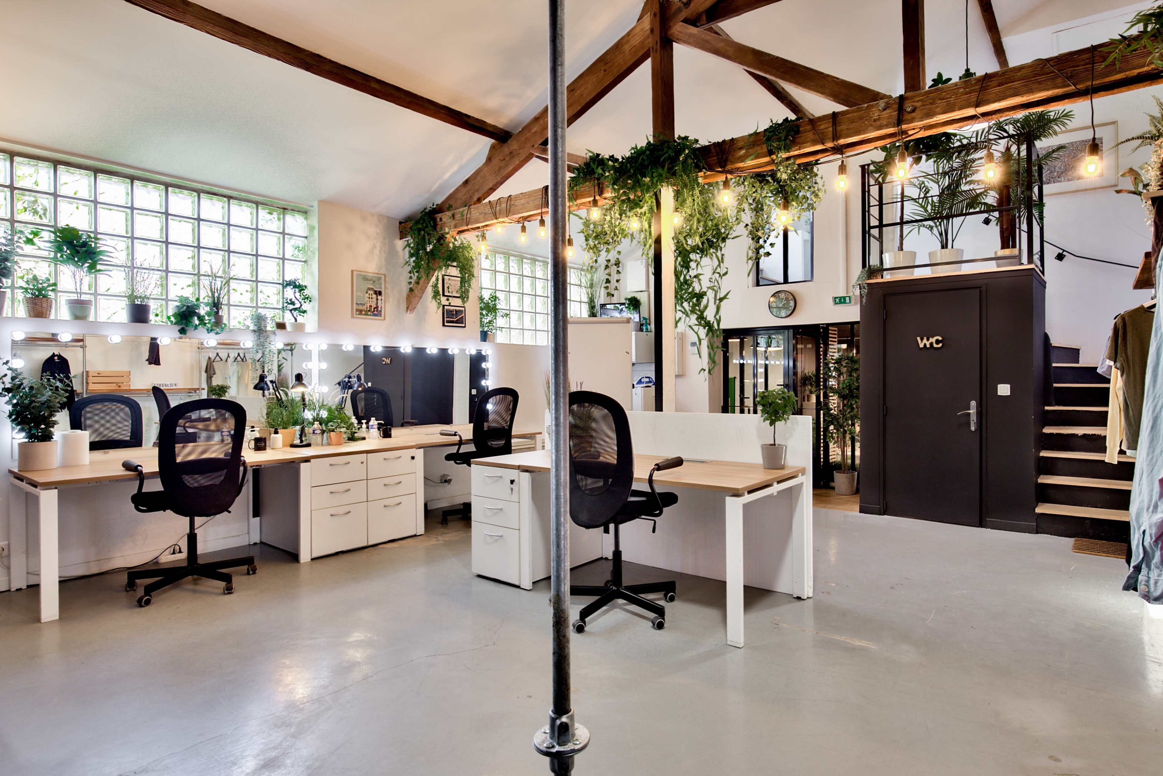 A spacious office interior featuring multiple workstations with plants, large windows, and an adjacent staircase.