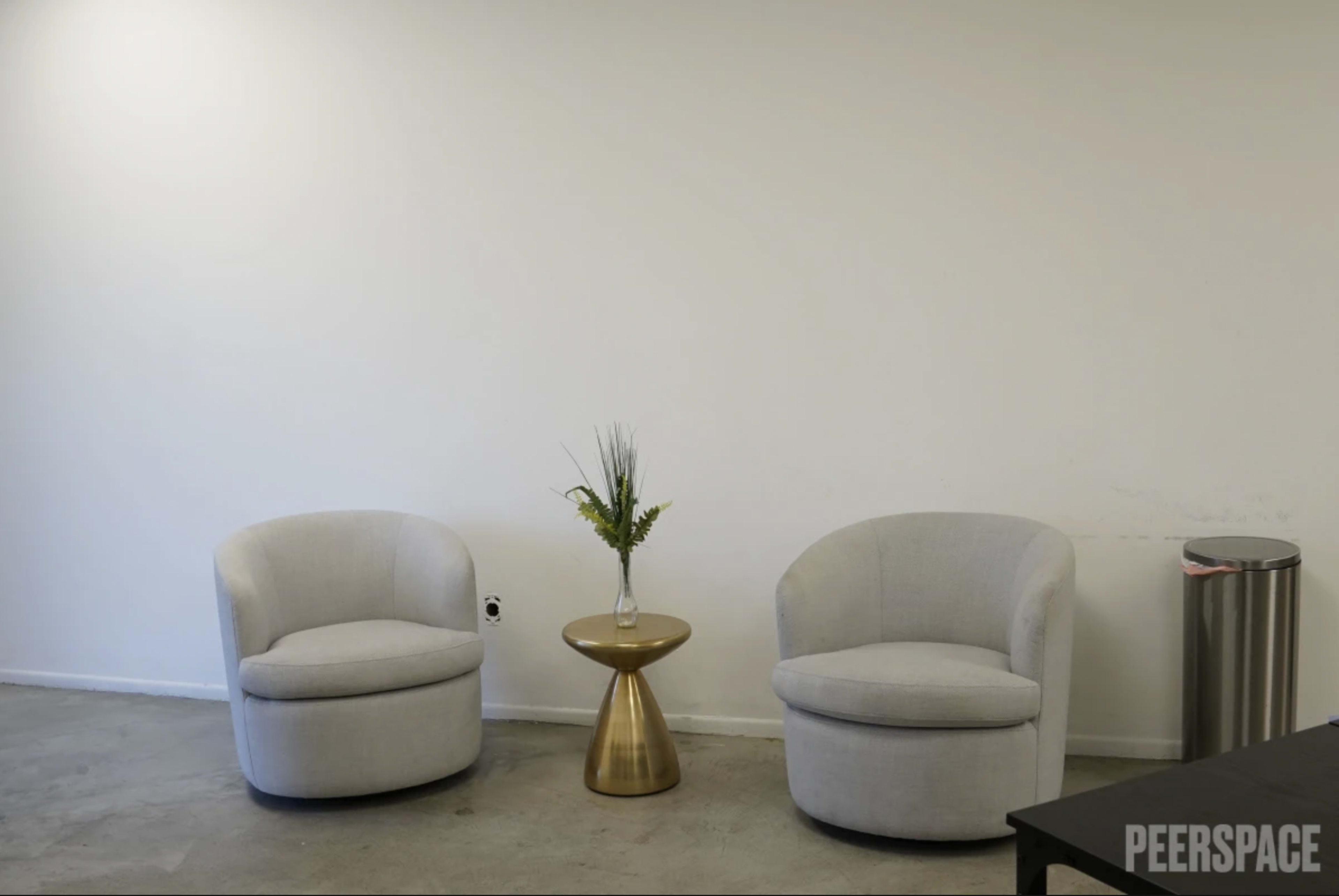 The image shows two light-colored armchairs flanking a small round gold side table with a vase of greenery, set against a plain white wall.