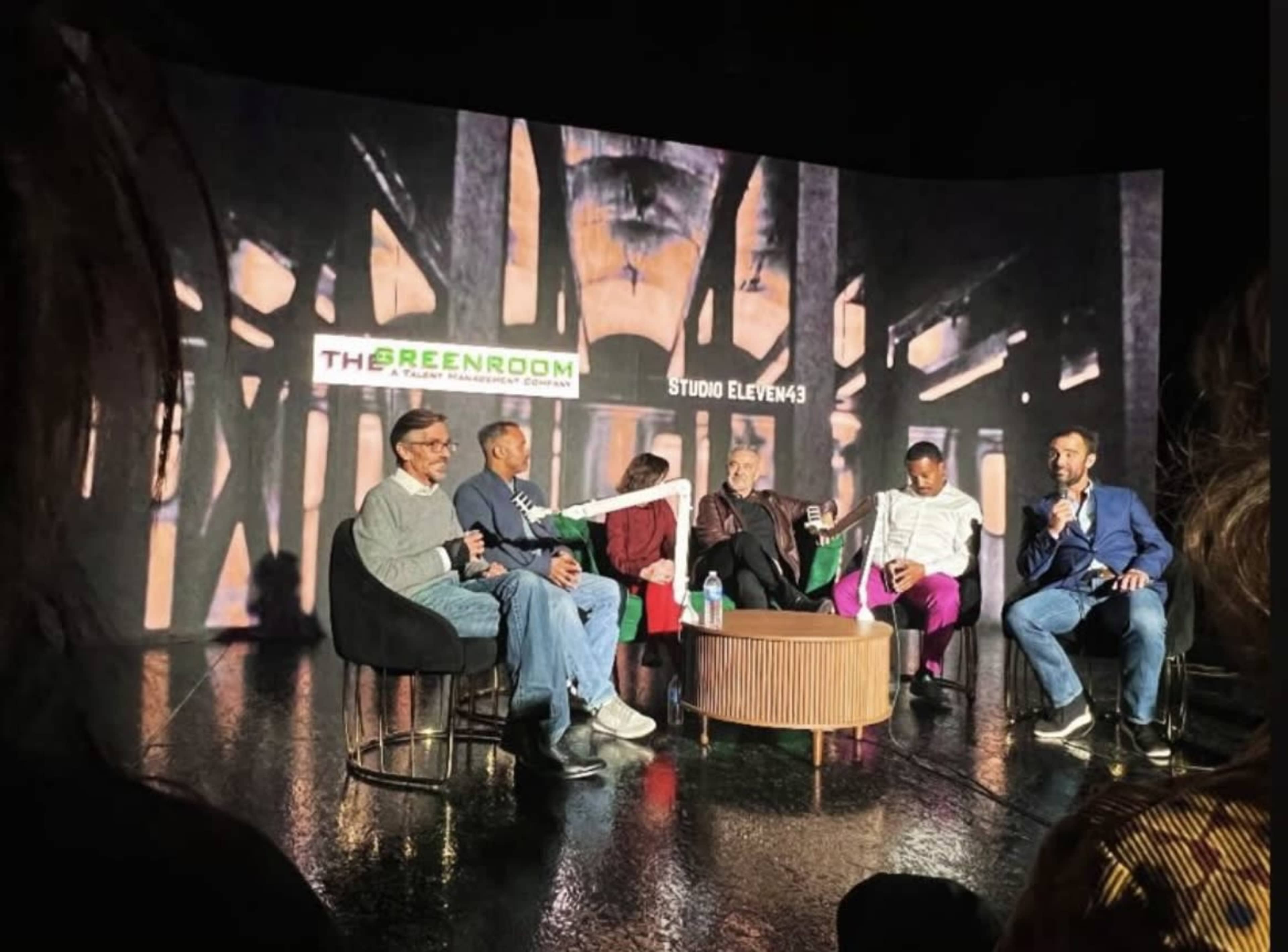 A panel discussion is taking place on stage, featuring five speakers seated in a semi-circle with a large screen displaying abstract visuals in the background.