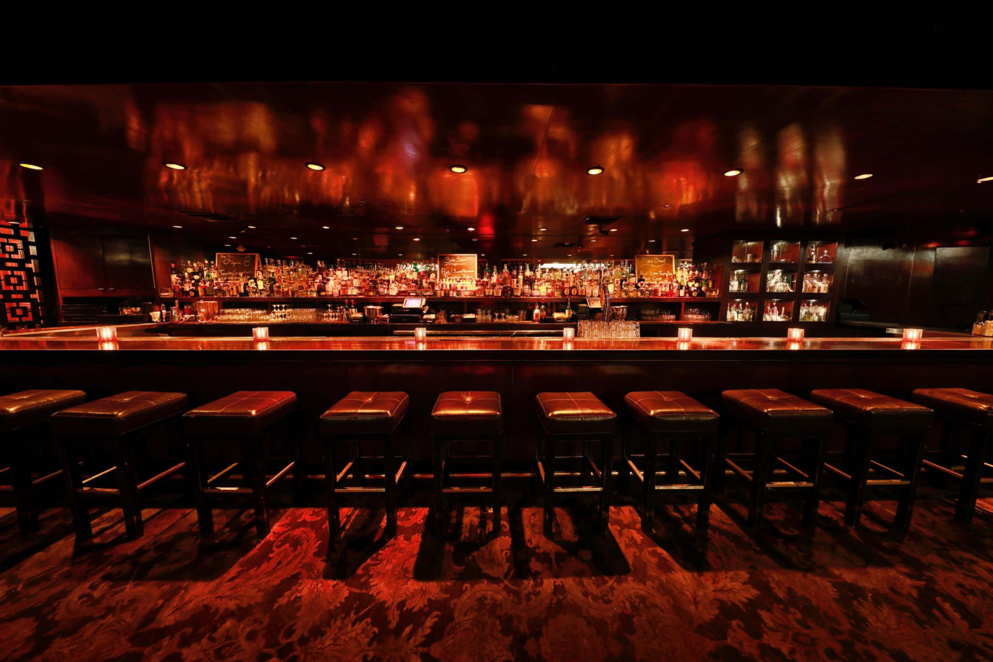 The image features a dimly lit bar with a long counter, illuminated by red lighting and lined with dark leather stools.