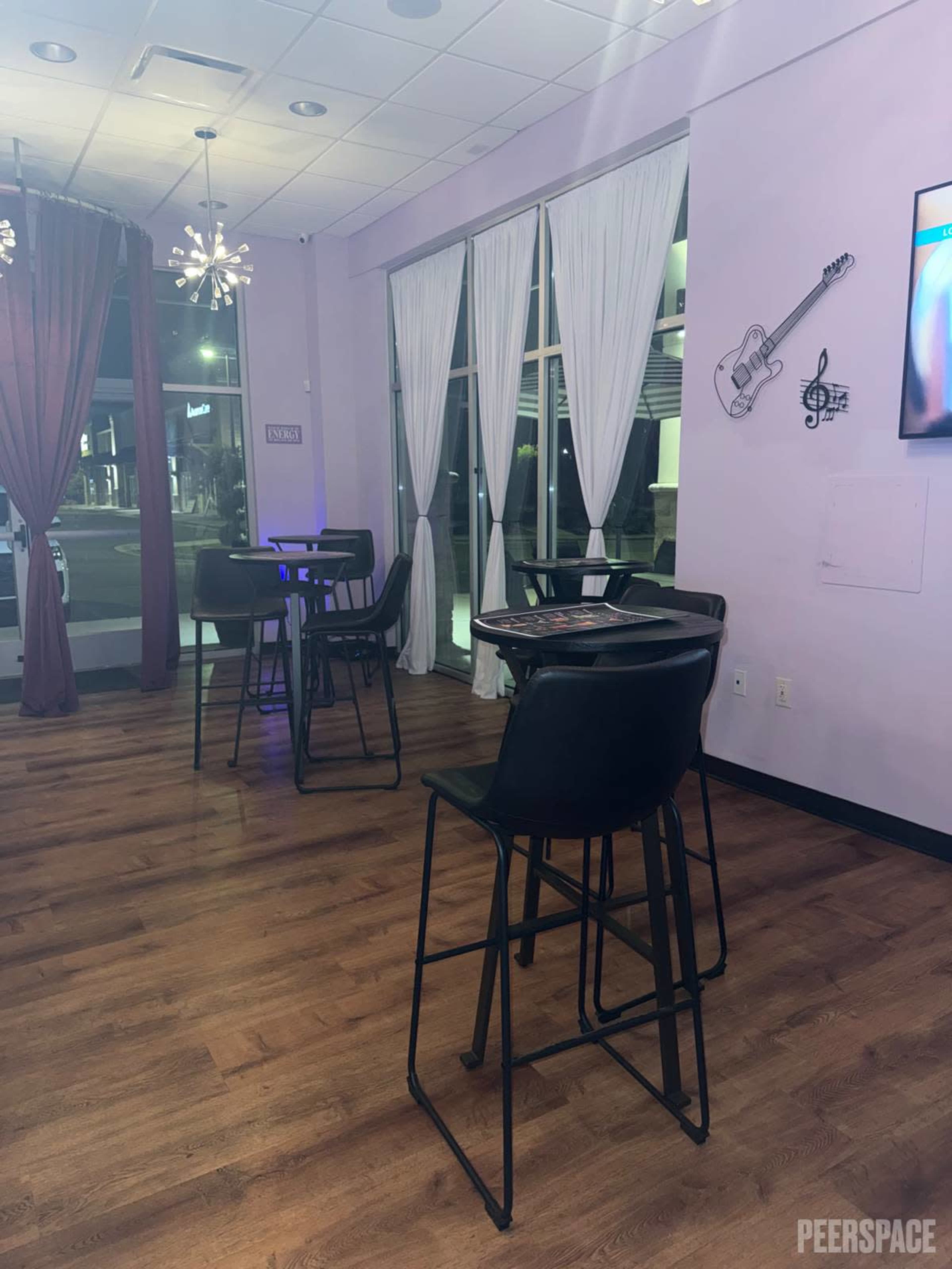 The image shows a brightly lit interior of a venue featuring several tall black tables and chairs against purple walls with decorative elements, including a guitar and musical notes.