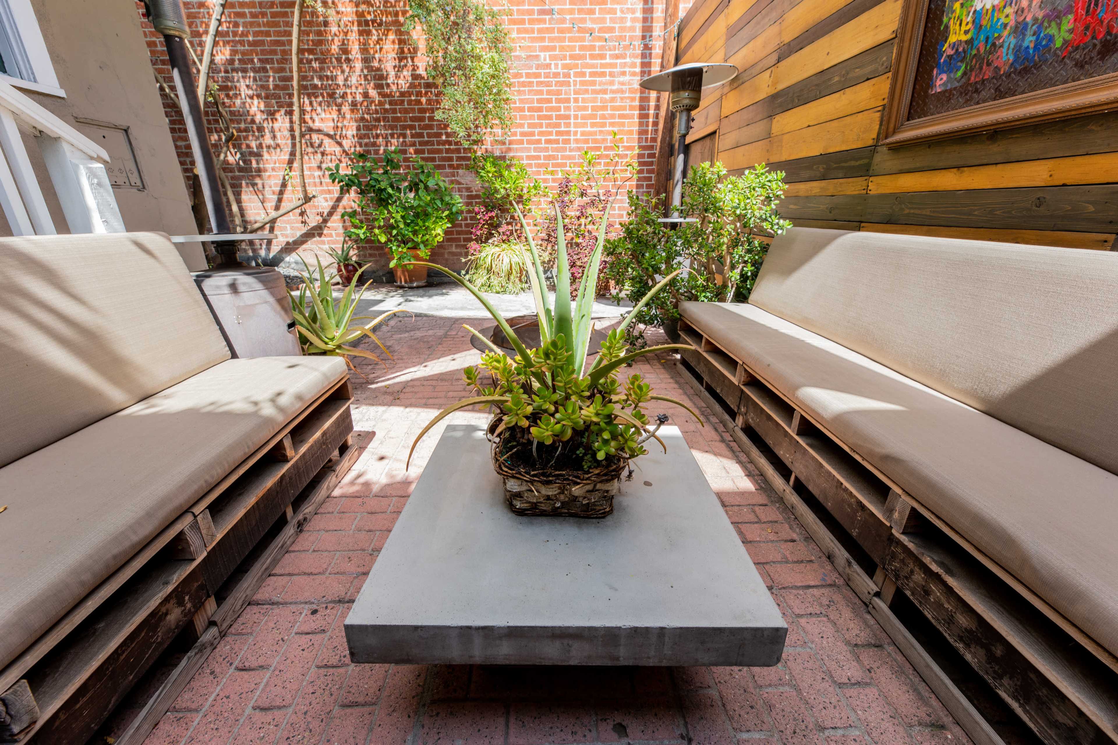 A small outdoor seating area features two benches made of wooden pallets facing a concrete table, surrounded by potted plants and brick walls.