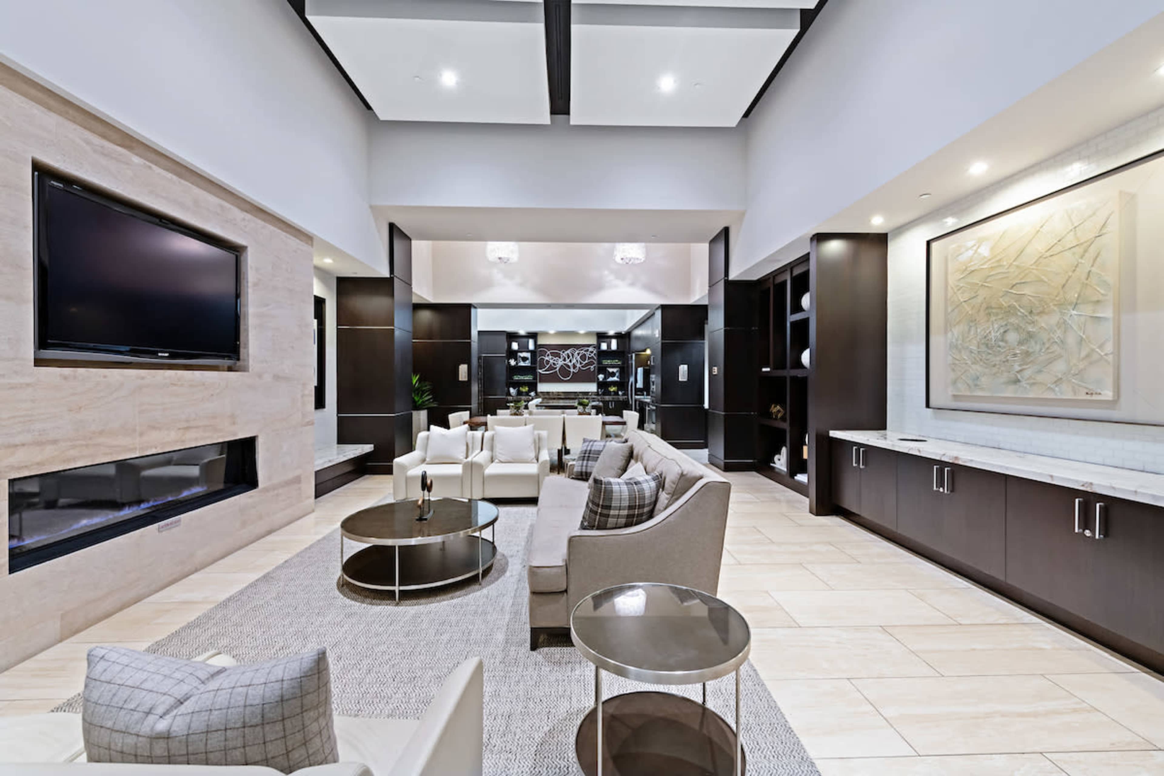 A modern living area features a large sectional sofa, glass coffee tables, and a mounted television against a neutral wall, with a dining area visible in the background.