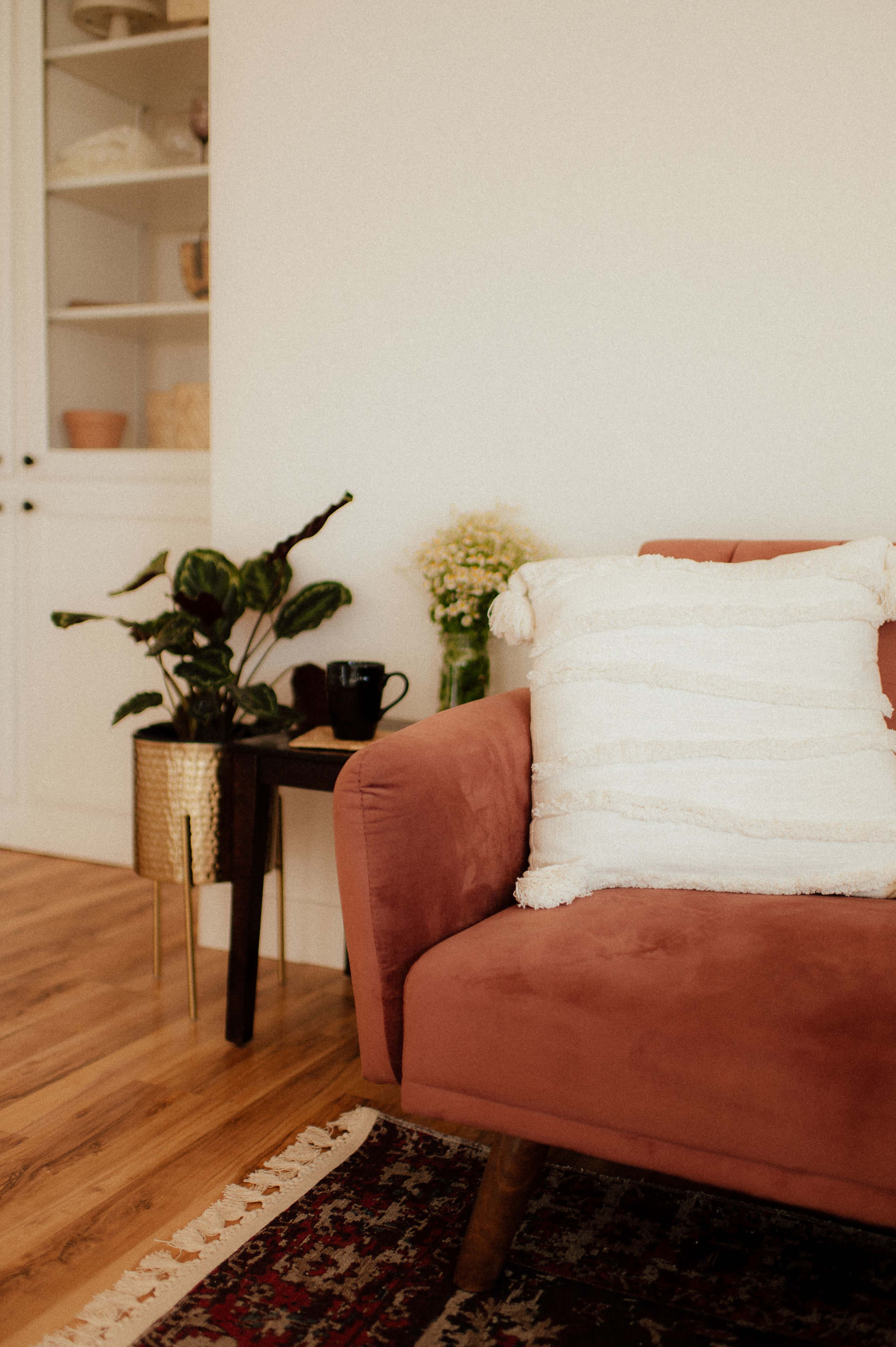 A pink sofa with a textured white pillow sits beside a small wooden table, accented by a plant and a black cup, in a bright room with wood flooring.