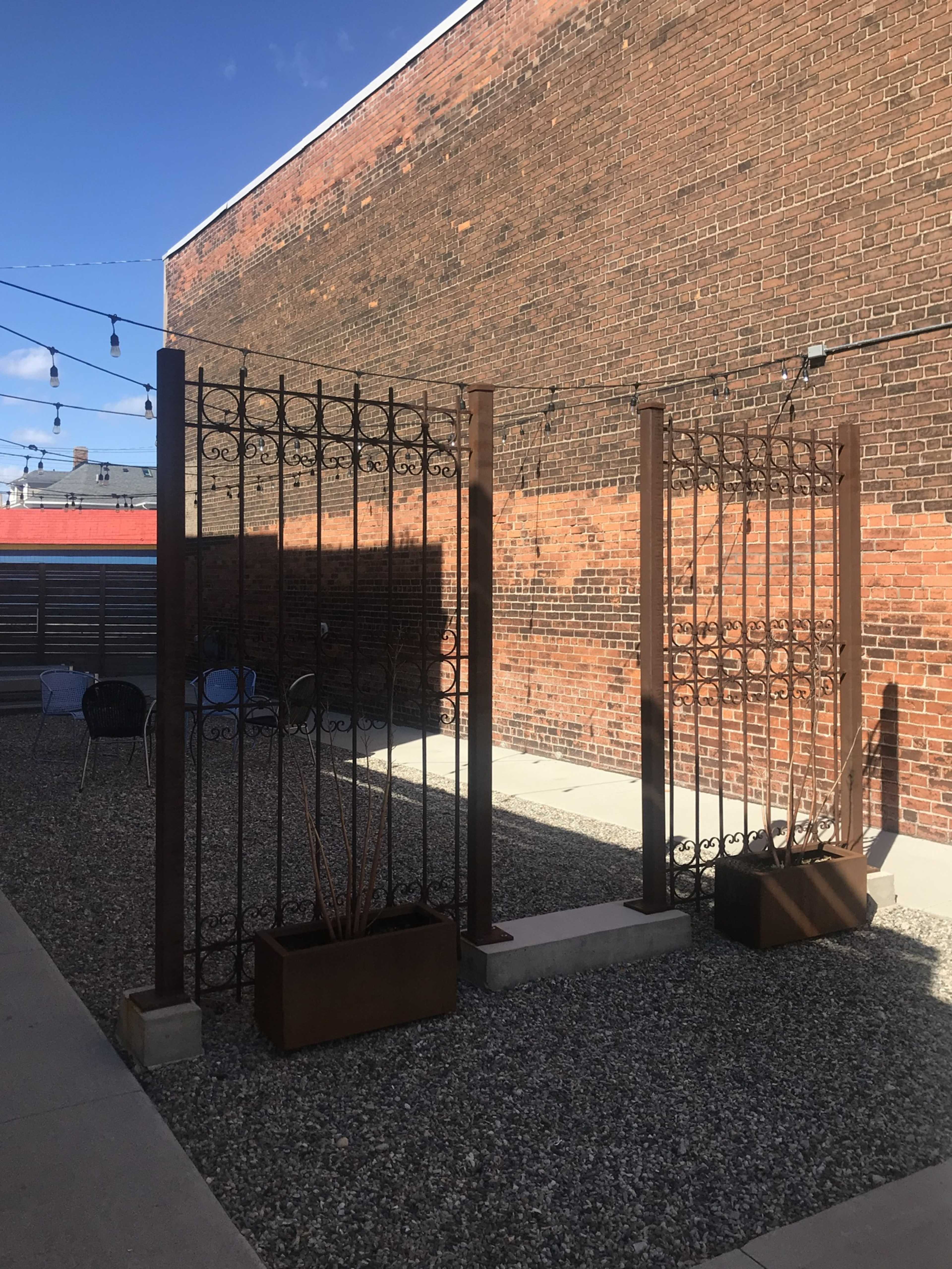 A gravel courtyard with two tall, ornate metal gates set against a brick wall, flanked by raised plant beds.