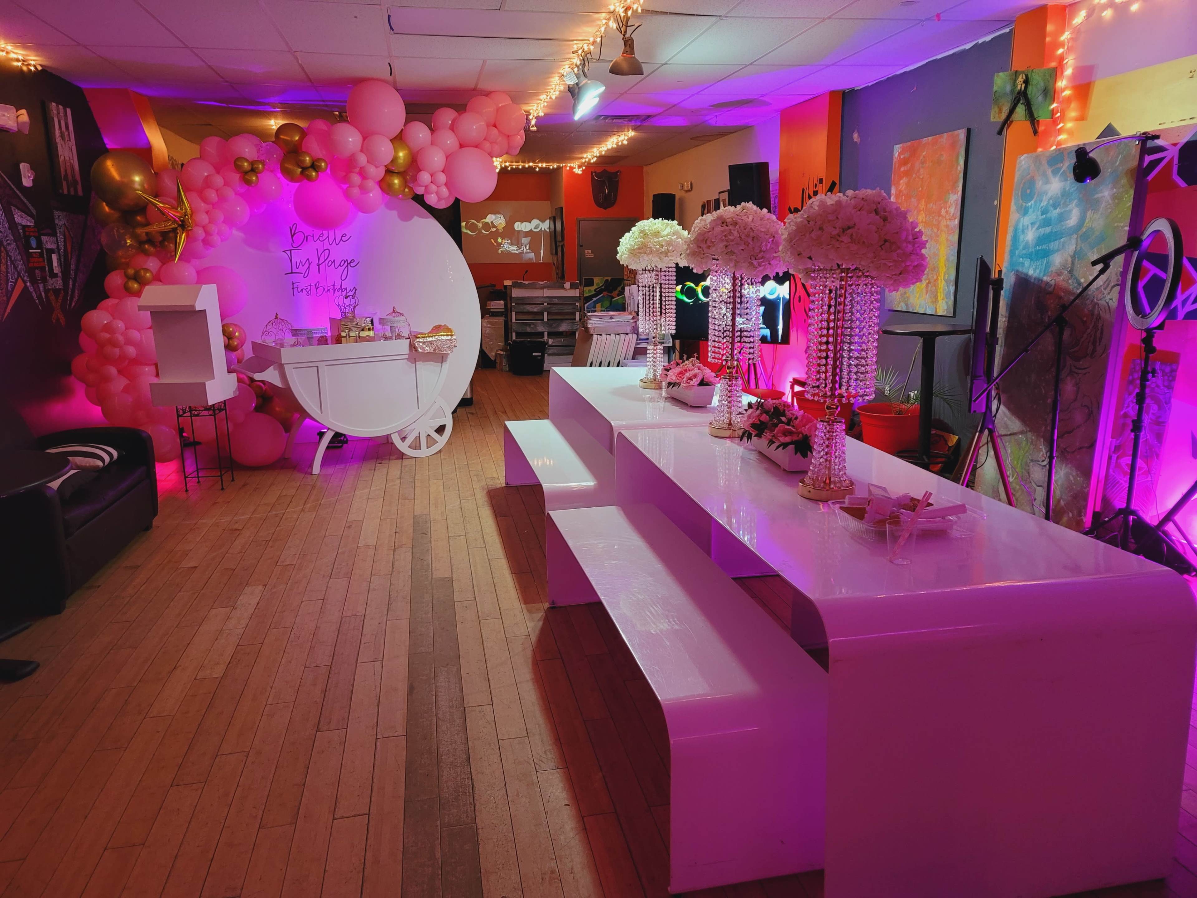 A brightly lit party venue with a decorative setup featuring tables, floral centerpieces, and balloon arches in a variety of colors.