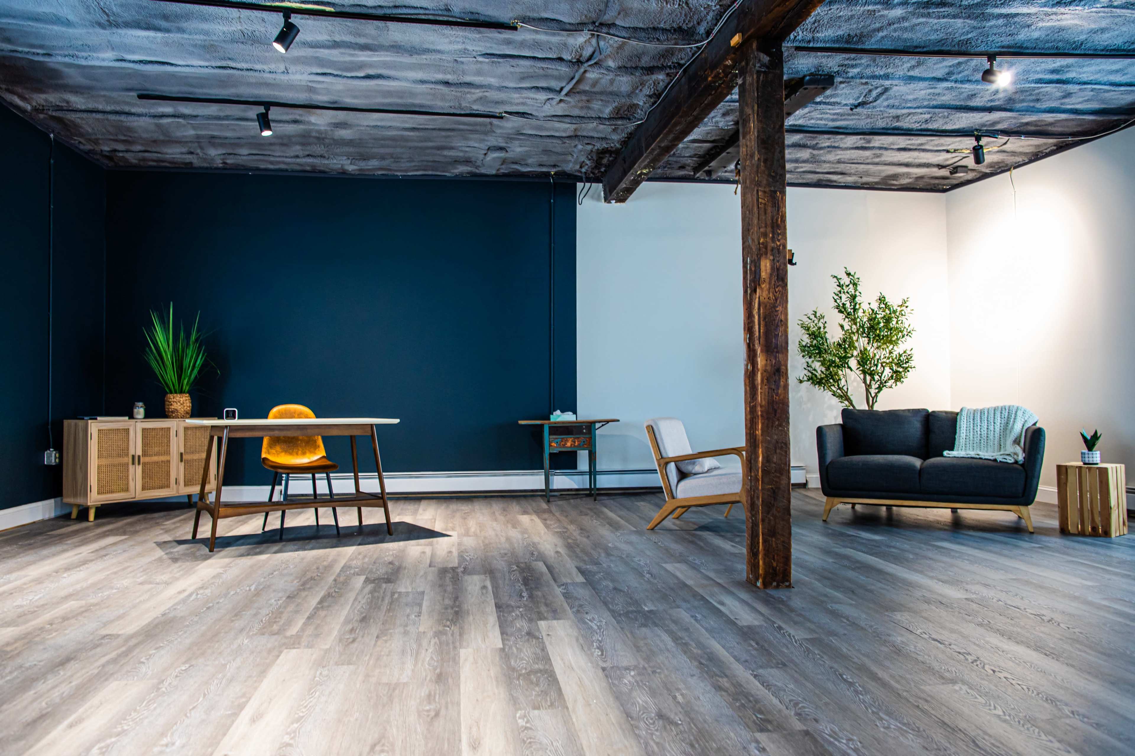 The image shows a spacious, empty room featuring a wooden floor, a dark blue wall, a wooden pillar, a desk with a chair, and a sofa with a plant nearby.